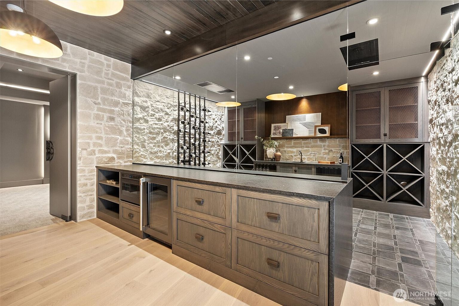 This sophisticated wine cellar features a custom-built bar area with dark wood cabinetry, a stone accent wall, and integrated wine storage racks. A large mirror reflects the space, adding depth and elegance, while the combination of natural stone, wood, and modern lighting creates a luxurious, moody atmosphere. The perspective is a wide-angle shot from the entrance, showcasing the seamless blend of functional storage and high-end design.