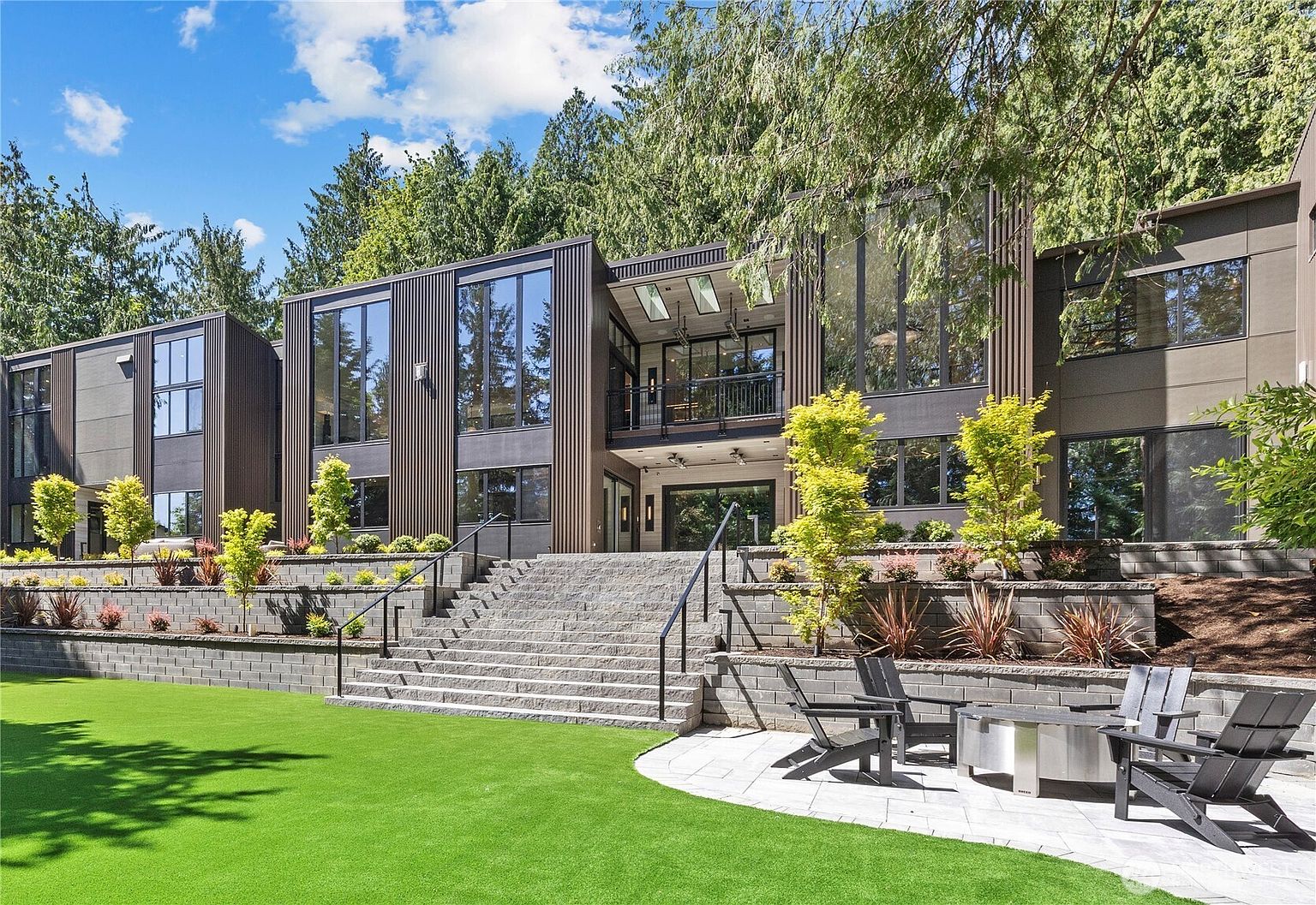 This striking modern residence features a multi-level architectural design with dark vertical siding, expansive floor-to-ceiling windows, and a prominent central balcony. A grand stone staircase leads from a manicured lawn and a paved patio area with a fire pit up to the main living level. The home is nestled against a lush, wooded backdrop, creating a sophisticated and serene outdoor living environment.