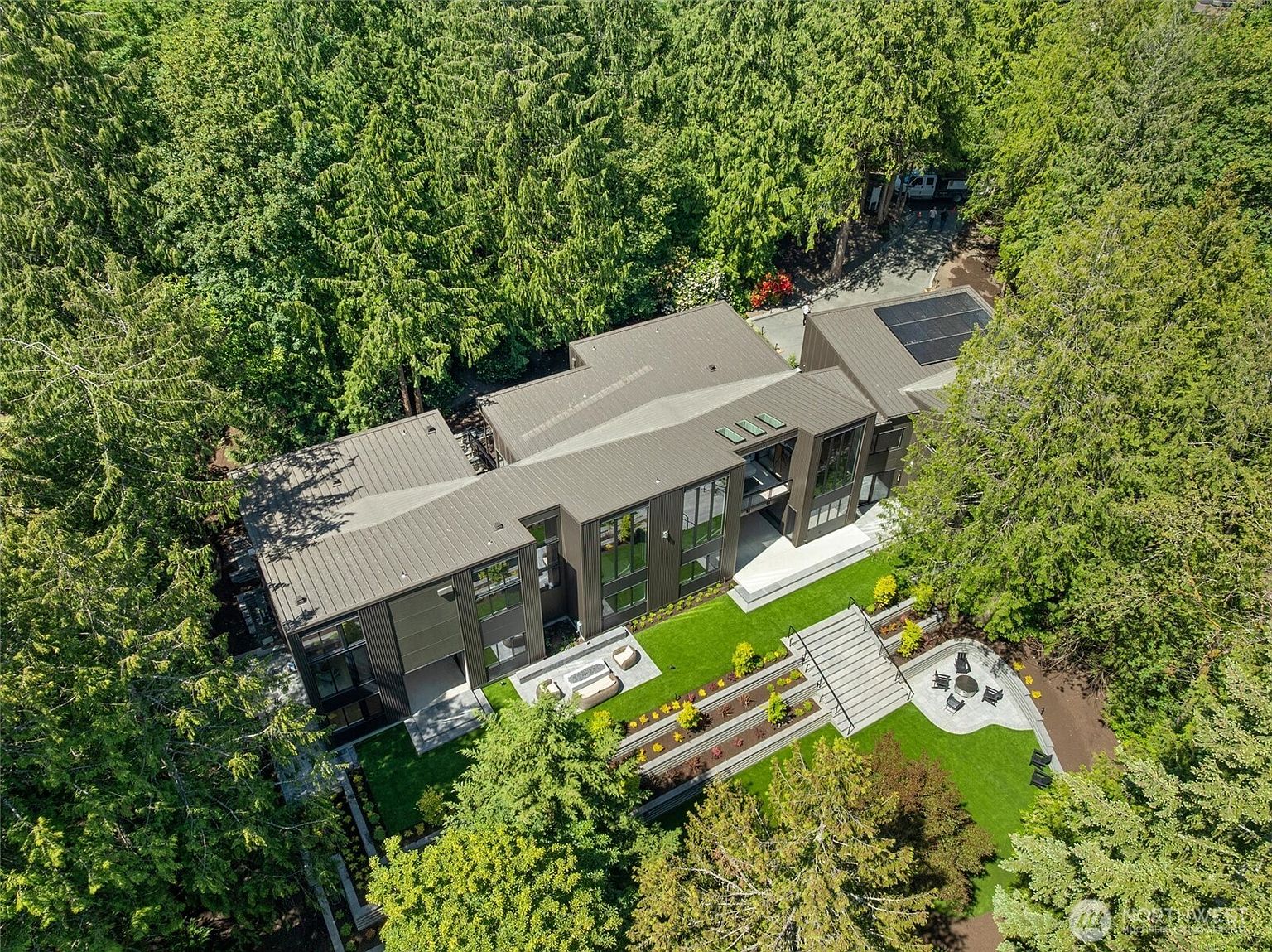 This high-angle aerial view captures a modern, multi-level architectural home nestled within a dense, lush forest. The property features a sleek, dark metal-clad exterior, expansive windows, and a meticulously landscaped backyard with tiered garden beds, a stone patio with a fire pit, and a grand staircase. The overall impression is one of sophisticated luxury integrated seamlessly into a serene, natural woodland setting.
