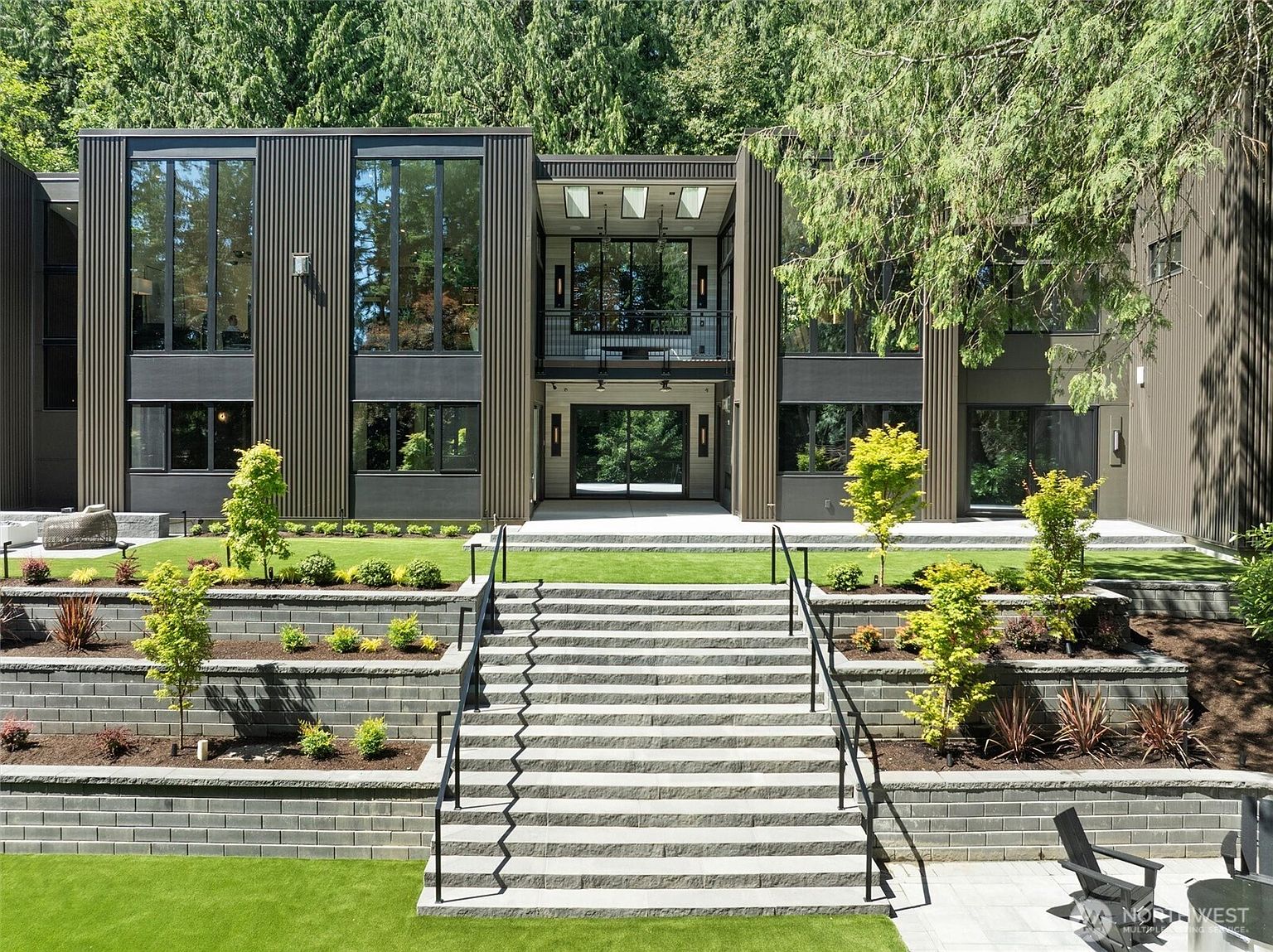 This striking modern residence features a bold, dark-toned vertical siding design with expansive floor-to-ceiling windows that create a seamless connection between the interior and the lush, forested surroundings. A grand, multi-tiered stone staircase serves as the focal point, leading up from a manicured lawn to a recessed, covered entryway and a second-story balcony. The architectural style is contemporary and sophisticated, emphasizing clean lines, geometric symmetry, and a harmonious integration with the natural landscape.