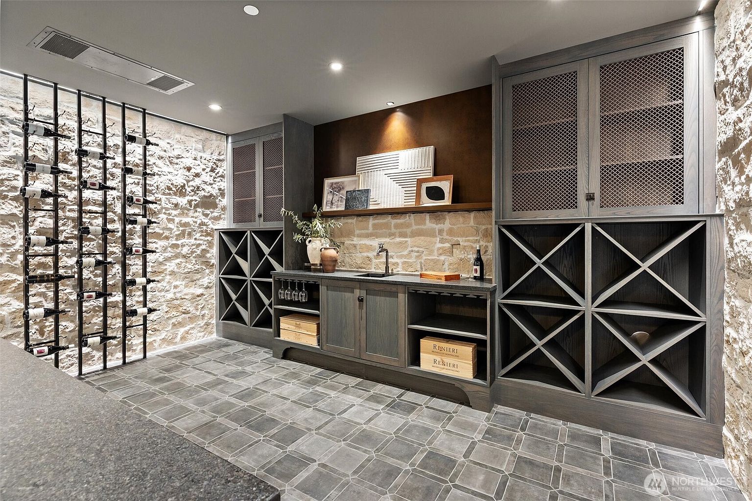 This sophisticated wine cellar features a rustic stone wall backdrop paired with custom dark-stained cabinetry and integrated metal wine racking. The space includes a functional wet bar with a sink, open shelving for storage crates, and elegant geometric floor tiling. The warm, recessed lighting creates an inviting and luxurious atmosphere perfect for wine storage and tasting.