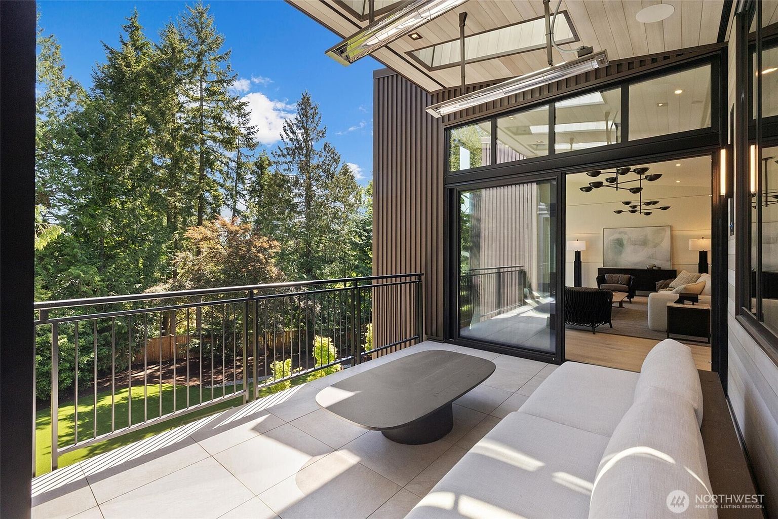 This modern, covered balcony features sleek tile flooring, a minimalist coffee table, and a comfortable outdoor sofa, creating a sophisticated lounge area. Large sliding glass doors provide a seamless transition to the interior living space, while the black metal railing overlooks a lush, tree-filled backyard. The architectural design emphasizes clean lines and a contemporary aesthetic, perfect for relaxing while enjoying the natural surroundings.