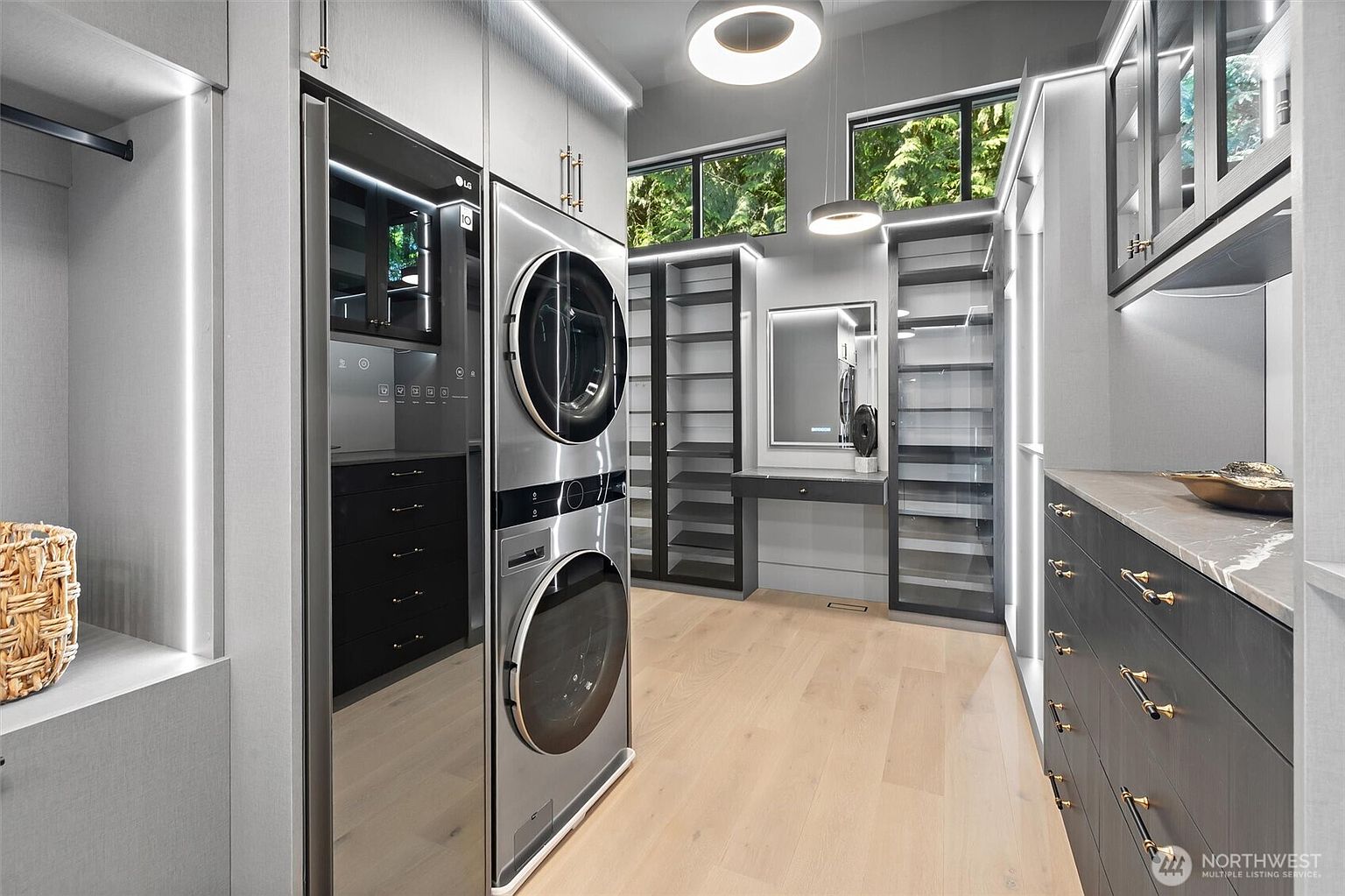 This sophisticated, modern laundry room features a sleek stacked LG washer and dryer unit integrated into custom cabinetry. The space is accented by warm wood flooring, recessed LED lighting, and a dedicated vanity area with a mirror, creating a high-end, functional aesthetic. Large windows allow natural light to filter in, highlighting the clean lines and premium finishes throughout the room.