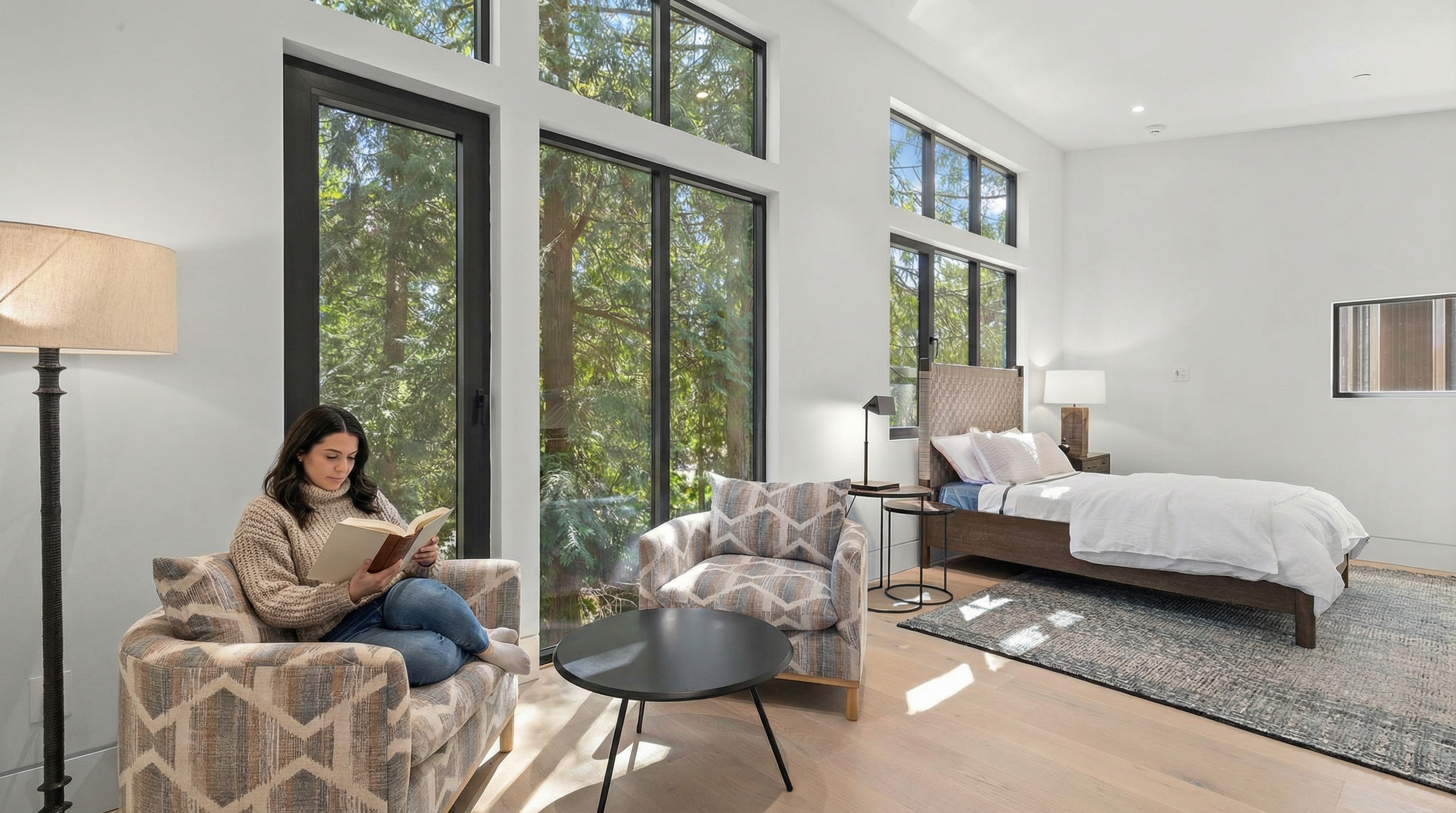 This modern, light-filled bedroom features floor-to-ceiling windows that offer a serene view of the surrounding forest. The room is furnished with a comfortable bed, a patterned armchair, and a small side table, creating a cozy and tranquil atmosphere. The neutral color palette and clean lines emphasize a contemporary design aesthetic.