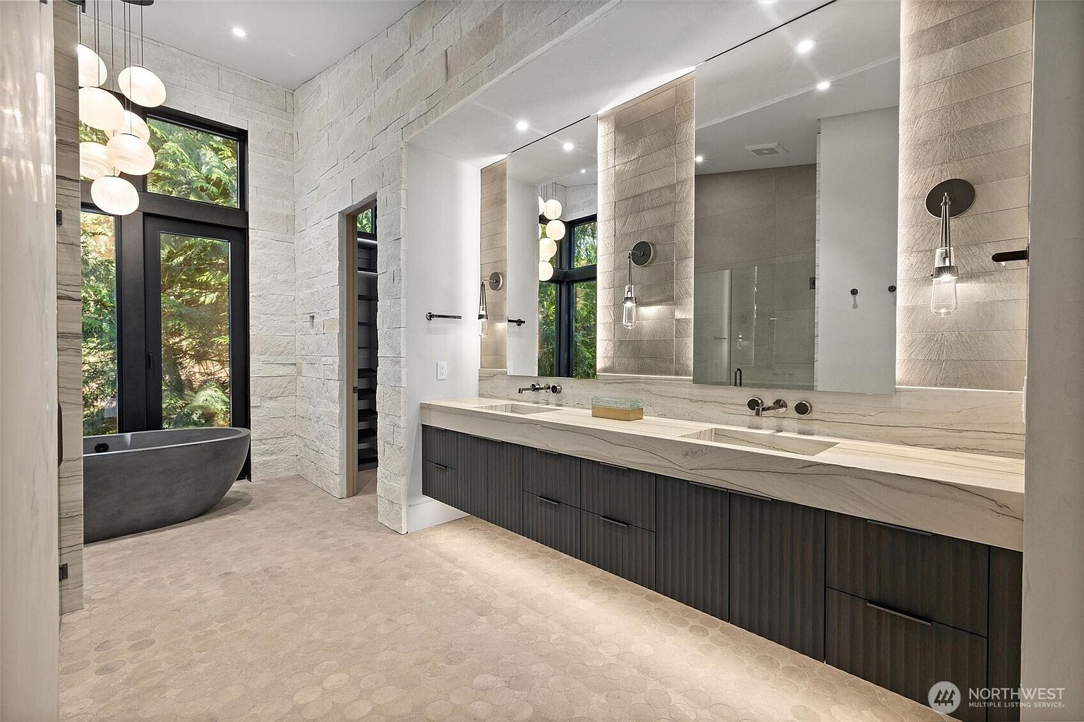 This luxurious primary bathroom features a sleek, floating double vanity with dark wood cabinetry and a striking stone countertop. A deep, charcoal-colored soaking tub sits elegantly near a floor-to-ceiling window, while textured stone walls and warm, integrated lighting create a sophisticated, spa-like atmosphere. The perspective captures the expansive layout, highlighting the seamless blend of natural materials and modern design elements.