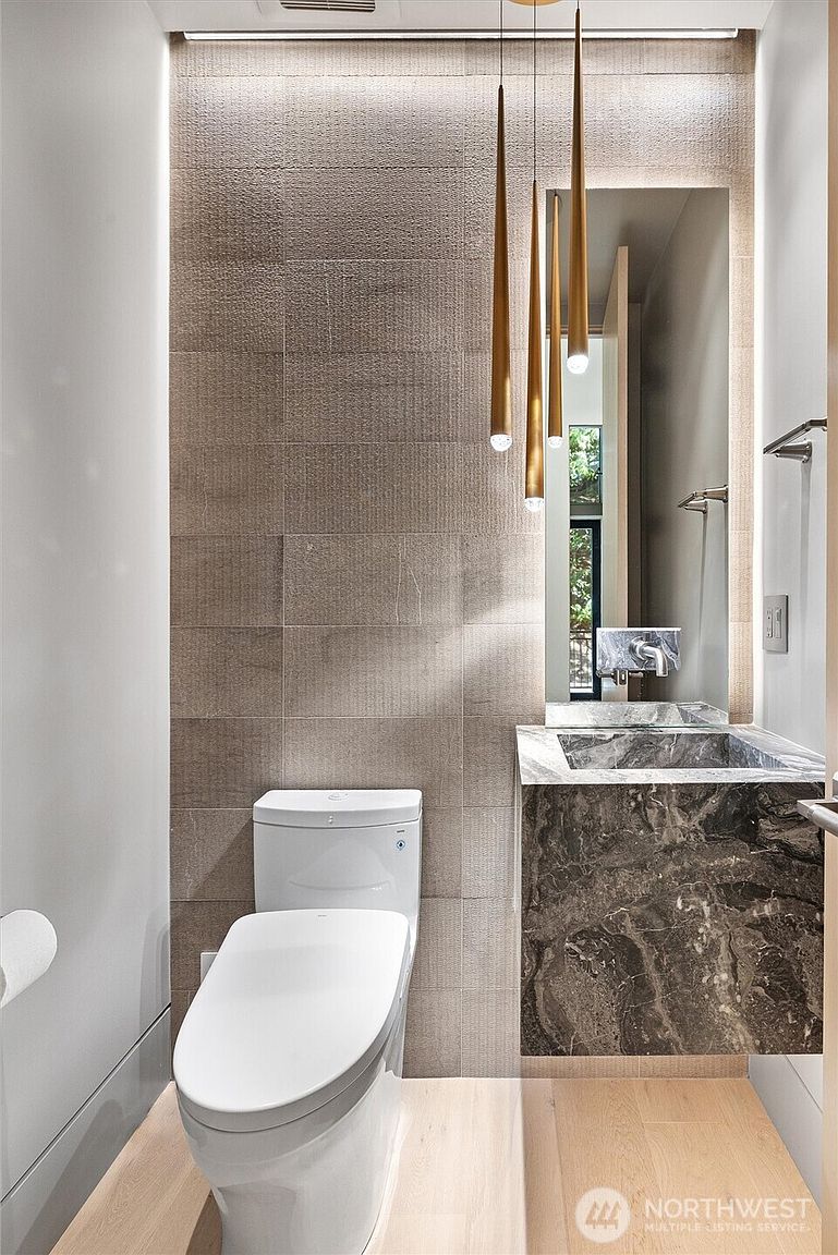 This modern powder room features a striking floating vanity crafted from dark, veined marble, paired with a sleek white toilet. The wall behind the vanity and toilet is clad in textured, neutral-toned stone tiles, accented by three elegant, slender gold pendant lights that create a warm, sophisticated ambiance. The space is finished with light wood flooring and a large, frameless mirror, offering a clean and luxurious aesthetic.