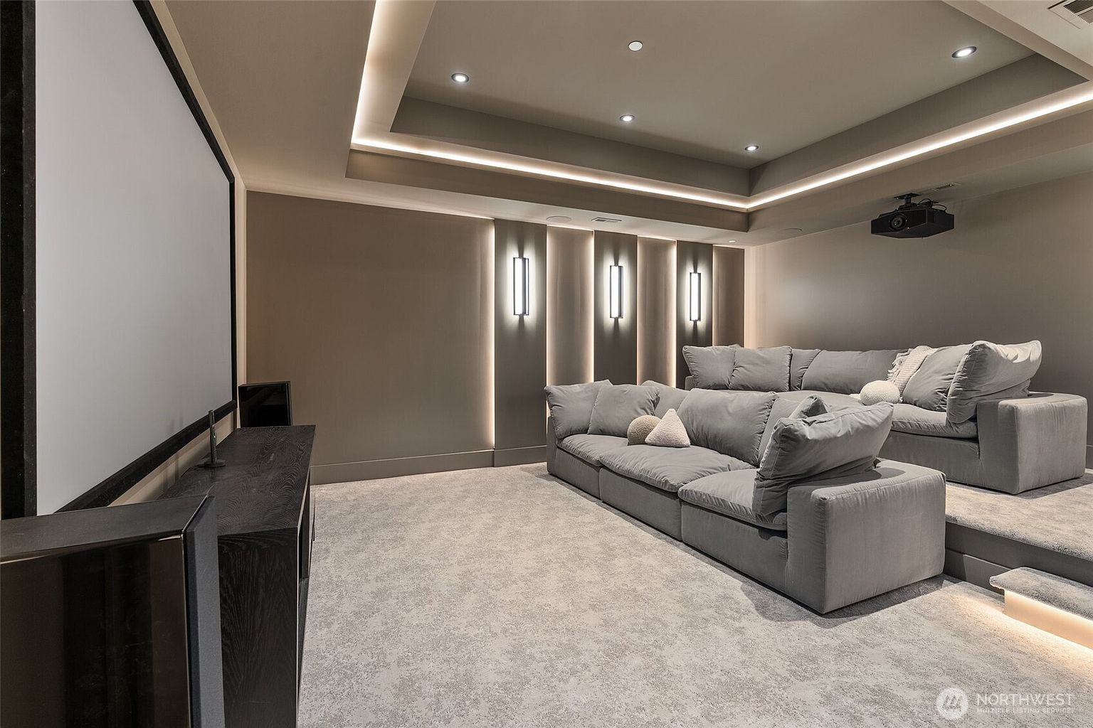 This sophisticated home theater features tiered seating with plush, oversized grey sectionals, creating a luxurious and comfortable viewing experience. The room is accented by modern recessed tray ceiling lighting and vertical wall sconces that provide a warm, ambient glow against the neutral-toned walls. A large projection screen and a dark wood media console complete the professional cinematic setup, making it an ideal space for entertainment.