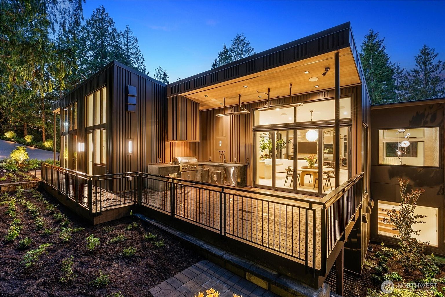 This stunning modern home features a spacious, elevated wooden deck complete with a built-in outdoor kitchen and stainless steel grill. The architectural design showcases sleek, dark vertical metal siding and expansive glass sliding doors that seamlessly blend indoor and outdoor living spaces. The scene is captured at dusk, with warm interior lighting creating an inviting and sophisticated atmosphere against the backdrop of a lush, forested landscape.