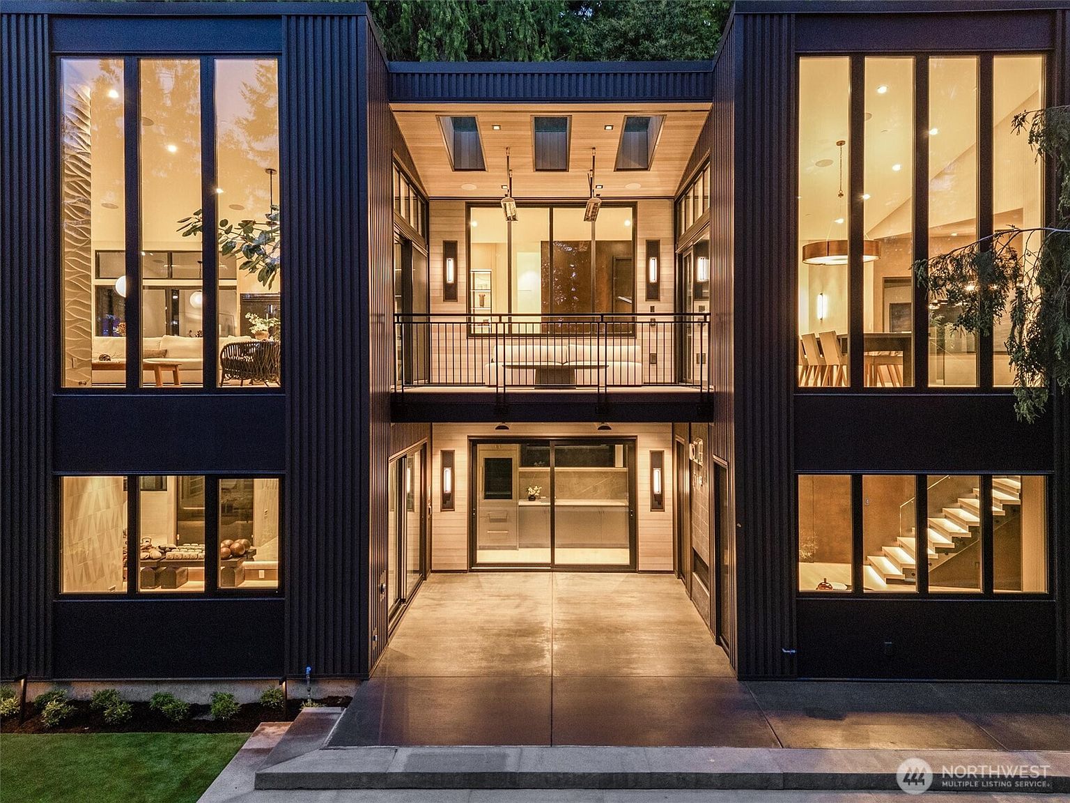This striking modern residence features a symmetrical, U-shaped architectural design with dark, vertical corrugated metal siding and expansive floor-to-ceiling windows. The central courtyard area includes a covered ground-level patio and a second-story balcony, both illuminated by warm, inviting interior lighting. The clean lines and contemporary aesthetic create a sophisticated, high-end look that emphasizes indoor-outdoor living.