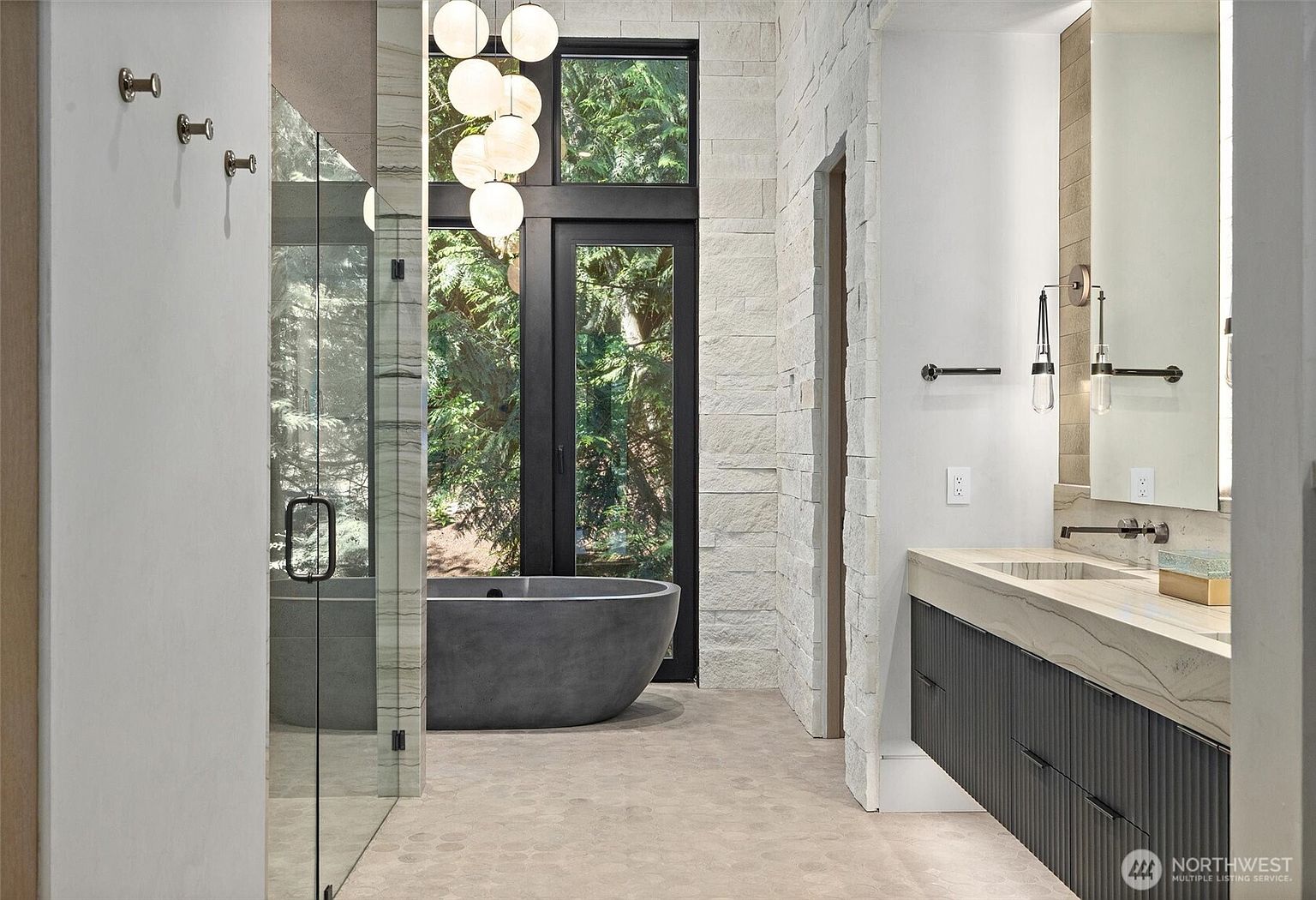 This luxurious primary bathroom features a striking charcoal-colored freestanding soaking tub positioned in front of a floor-to-ceiling window that overlooks a lush, wooded landscape. The space is anchored by a modern floating vanity with a textured dark cabinet and a light stone countertop, complemented by elegant globe pendant lighting. The combination of natural stone wall textures, glass shower enclosures, and organic views creates a serene, spa-like atmosphere.