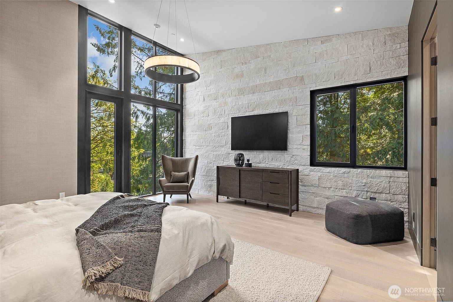 This spacious primary bedroom features a striking floor-to-ceiling stone accent wall and expansive windows that invite natural light and forest views. The room is furnished with a plush bed, a modern dark wood dresser, and a comfortable armchair, all set against light-toned hardwood flooring. The overall aesthetic is a sophisticated blend of contemporary design and natural textures, creating a serene and luxurious retreat.
