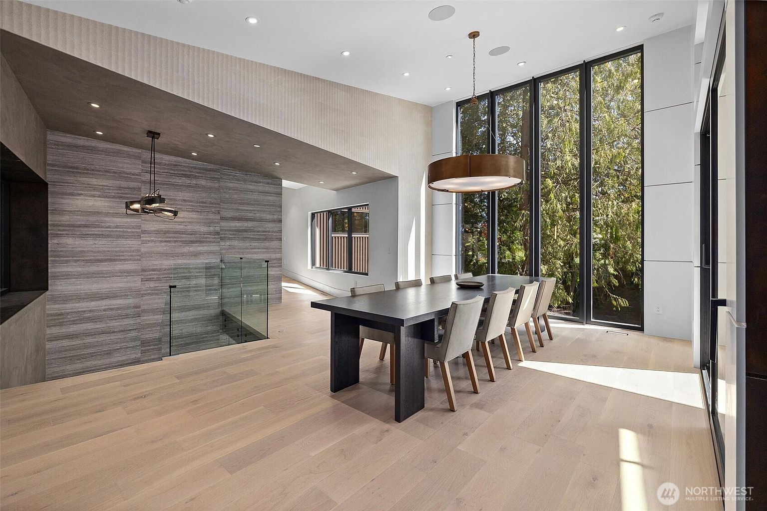 This sophisticated dining room features a long, dark wood table surrounded by eight upholstered chairs, set against a backdrop of floor-to-ceiling windows that offer a lush, wooded view. The space is defined by a striking contrast between the warm, light-toned hardwood floors and the textured, grey-toned feature wall that leads toward a glass-railed staircase. A large, modern drum pendant light hangs above the table, enhancing the room's contemporary and airy architectural aesthetic.