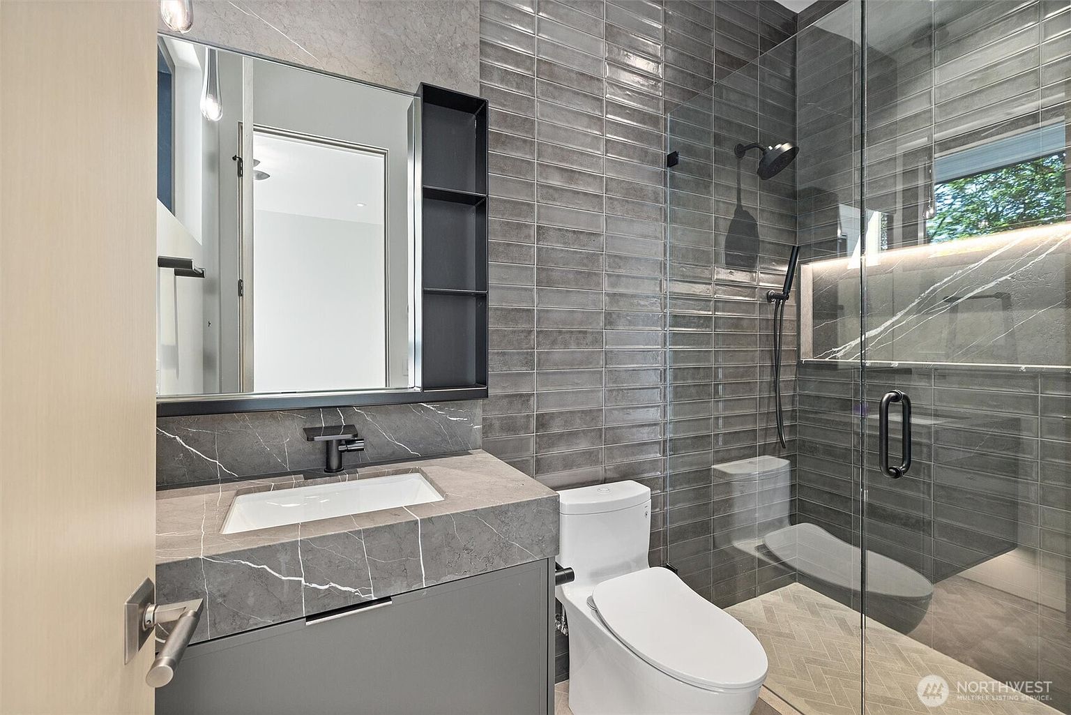 This modern, compact bathroom features a sleek gray vanity with a marble countertop and an integrated sink, paired with a contemporary mirror and shelving unit. The walk-in shower is enclosed by a glass partition and showcases textured gray wall tiles and a herringbone-patterned floor. The overall aesthetic is sophisticated and minimalist, utilizing a monochromatic color palette to create a clean, high-end feel.