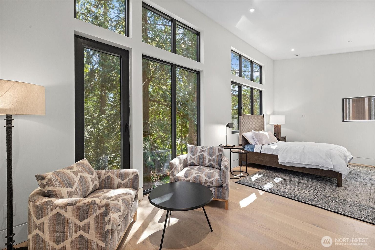 This modern, light-filled bedroom features floor-to-ceiling windows that offer a serene view of the surrounding forest. The room is furnished with a comfortable bed, a patterned armchair, and a small side table, creating a cozy and tranquil atmosphere. The neutral color palette and clean lines emphasize a contemporary design aesthetic.