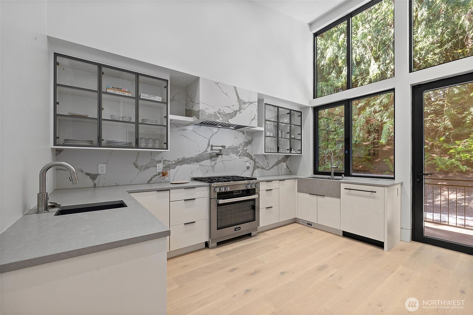 This modern, high-ceilinged kitchen features sleek white cabinetry paired with striking marble-patterned backsplash and countertops. The space is equipped with premium stainless steel appliances, including a professional-grade range and a farmhouse-style sink, while large windows provide an abundance of natural light and a serene view of the surrounding forest. The overall aesthetic is clean, sophisticated, and airy, emphasizing a seamless blend of luxury and nature.