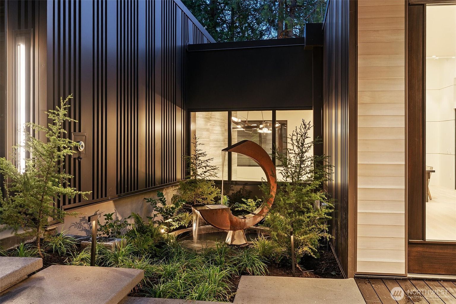 This sophisticated entryway features a striking, modern metal sculpture fountain as its centerpiece, set within a lush, landscaped garden bed. The architectural design utilizes vertical black metal siding and warm wood accents, creating a high-contrast, contemporary aesthetic. The scene is captured from a low angle, emphasizing the clean lines of the concrete steps and the inviting, illuminated transition into the home.