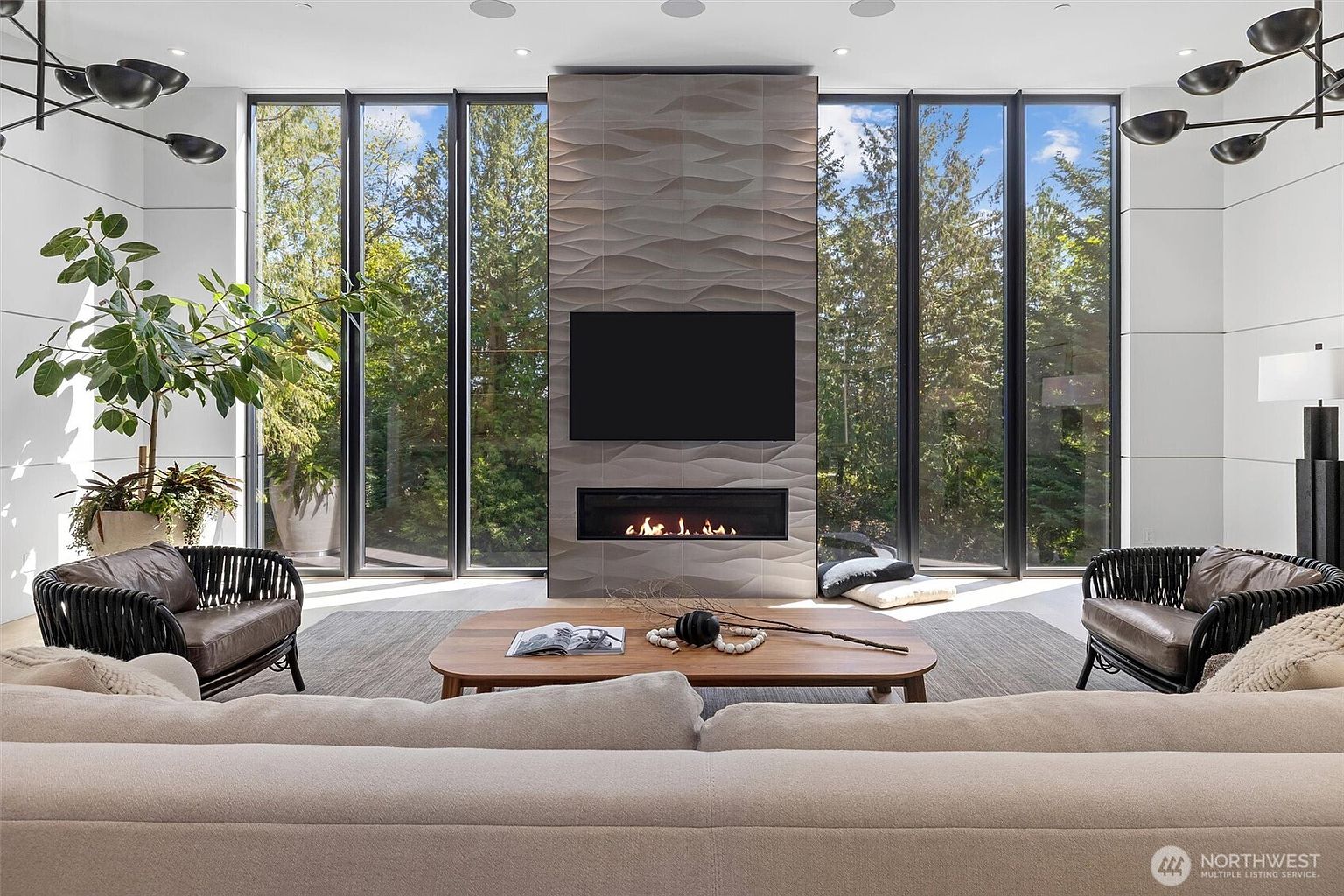 This sophisticated living room features a striking floor-to-ceiling fireplace clad in textured, wave-patterned stone, serving as the central focal point. Flanked by expansive, full-height windows that invite lush forest views inside, the space is anchored by a plush, neutral-toned sectional and two modern, dark-wicker accent chairs. The minimalist aesthetic is complemented by a warm wood coffee table and elegant, contemporary light fixtures, creating a serene and luxurious atmosphere.
