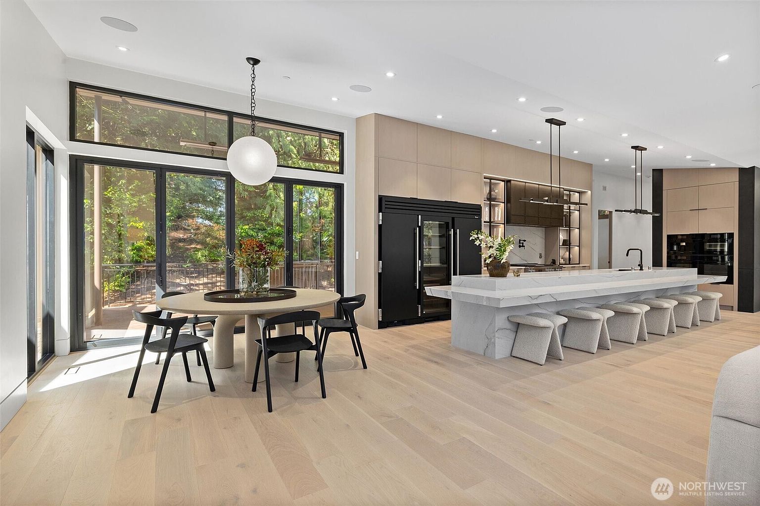 This expansive, modern open-concept kitchen and dining area features light hardwood flooring, a massive marble-topped island with seating for six, and sleek, floor-to-ceiling cabinetry. A round dining table sits adjacent to large, black-framed glass doors that provide a seamless transition to the outdoor deck, bathing the space in natural light. The design aesthetic is minimalist and sophisticated, emphasizing clean lines and high-end finishes.