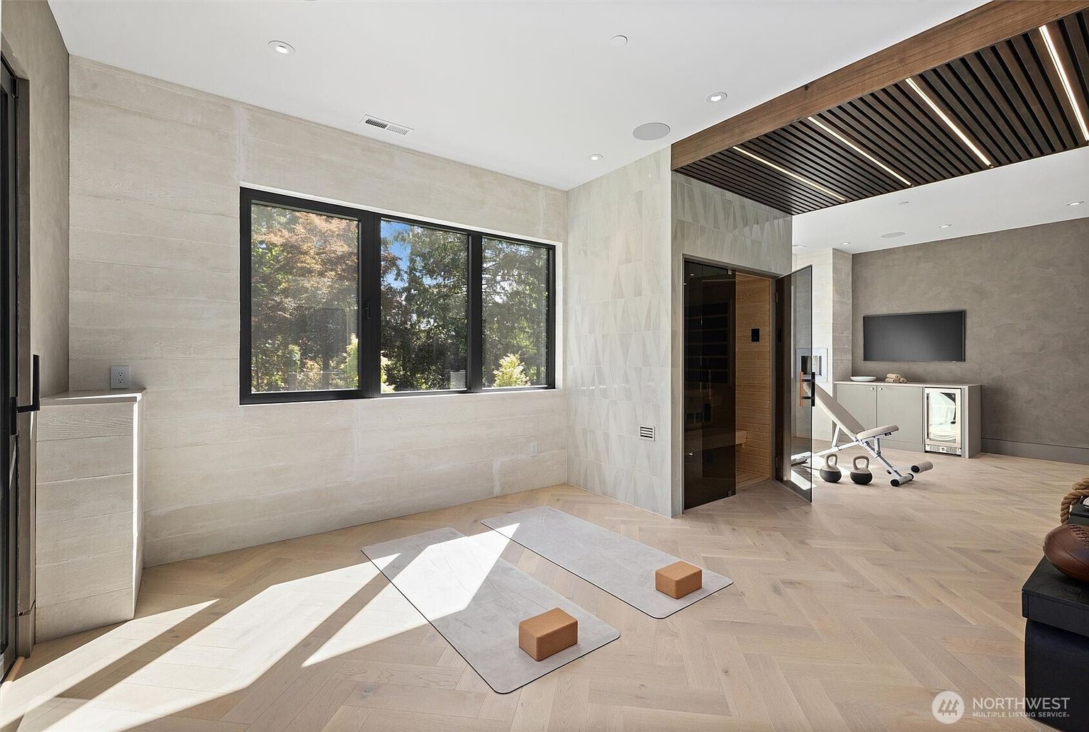This sophisticated home gym features a serene yoga area with two mats and blocks set against a light-toned, textured wall and herringbone wood flooring. The space seamlessly transitions into a fitness zone equipped with a weight bench, a sauna with a glass door, and a refreshment station with a mini-fridge and wall-mounted television. The room is bathed in natural light from large windows, creating a tranquil yet functional environment for wellness and exercise.