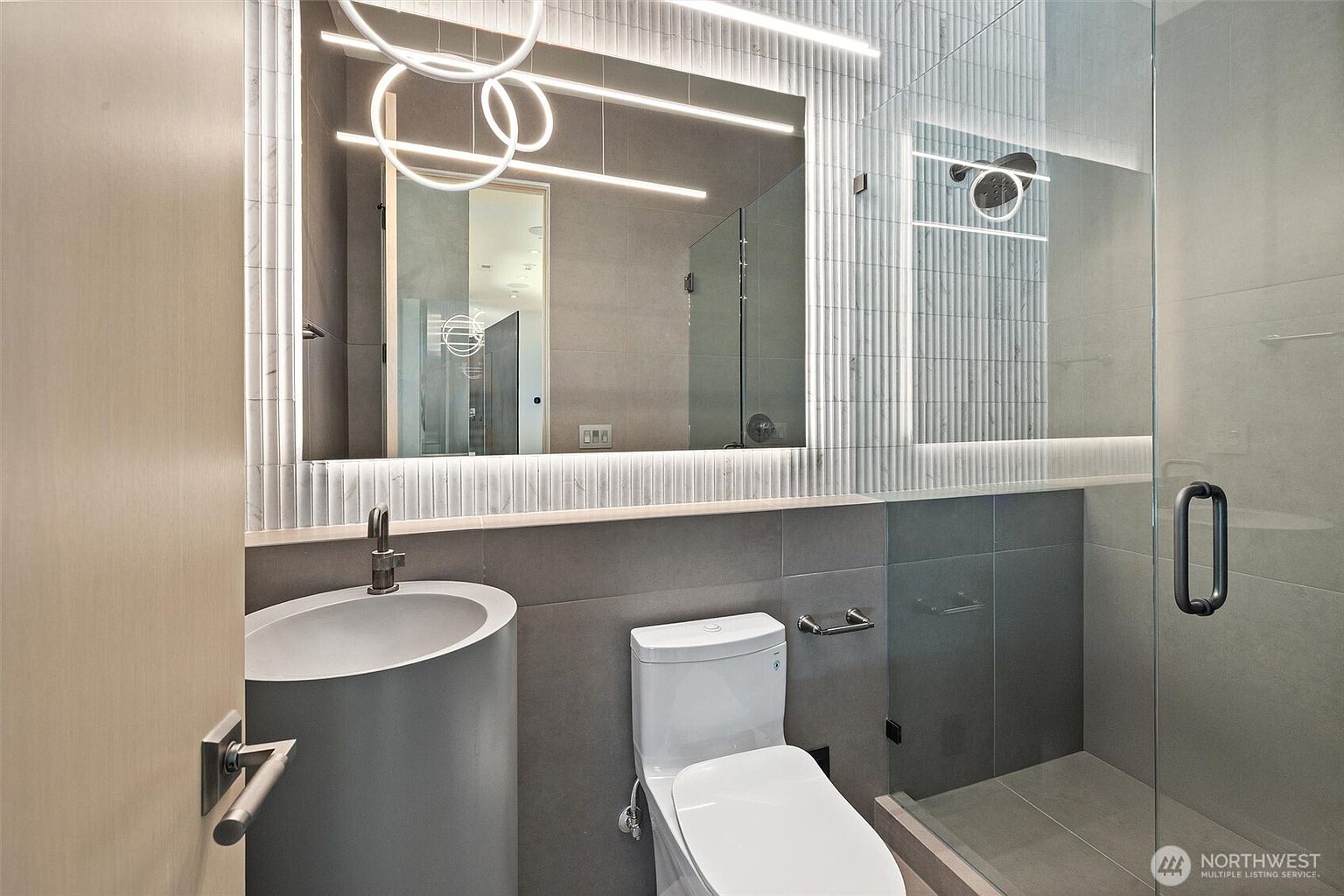 This modern, compact bathroom features a sleek cylindrical pedestal sink and a minimalist white toilet set against large-format grey wall tiles. A large, illuminated mirror with integrated linear lighting serves as the focal point, complemented by a glass-enclosed walk-in shower. The space exudes a contemporary, high-end aesthetic with clean lines and sophisticated lighting design.