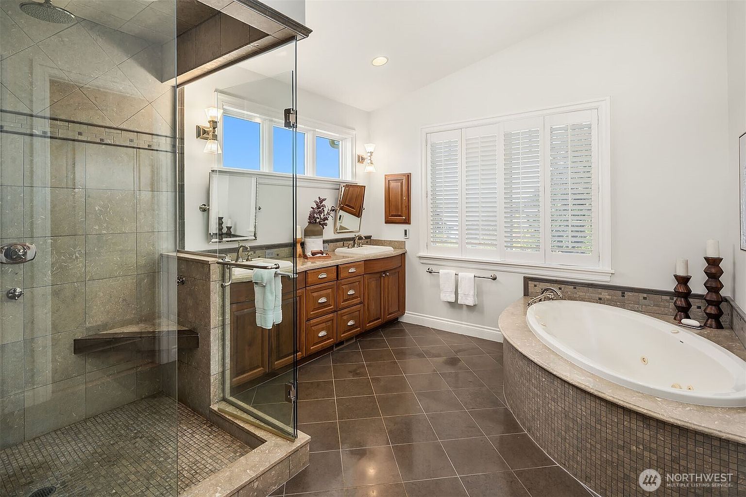 This spacious primary bathroom features a luxurious soaking tub with a tiled surround, a large glass-enclosed walk-in shower, and a double vanity with rich wood cabinetry. The room is brightened by natural light streaming through plantation shutters and windows above the vanity, creating an elegant and serene atmosphere. The dark tile flooring provides a sophisticated contrast to the light walls, emphasizing the high-end finishes of the space.