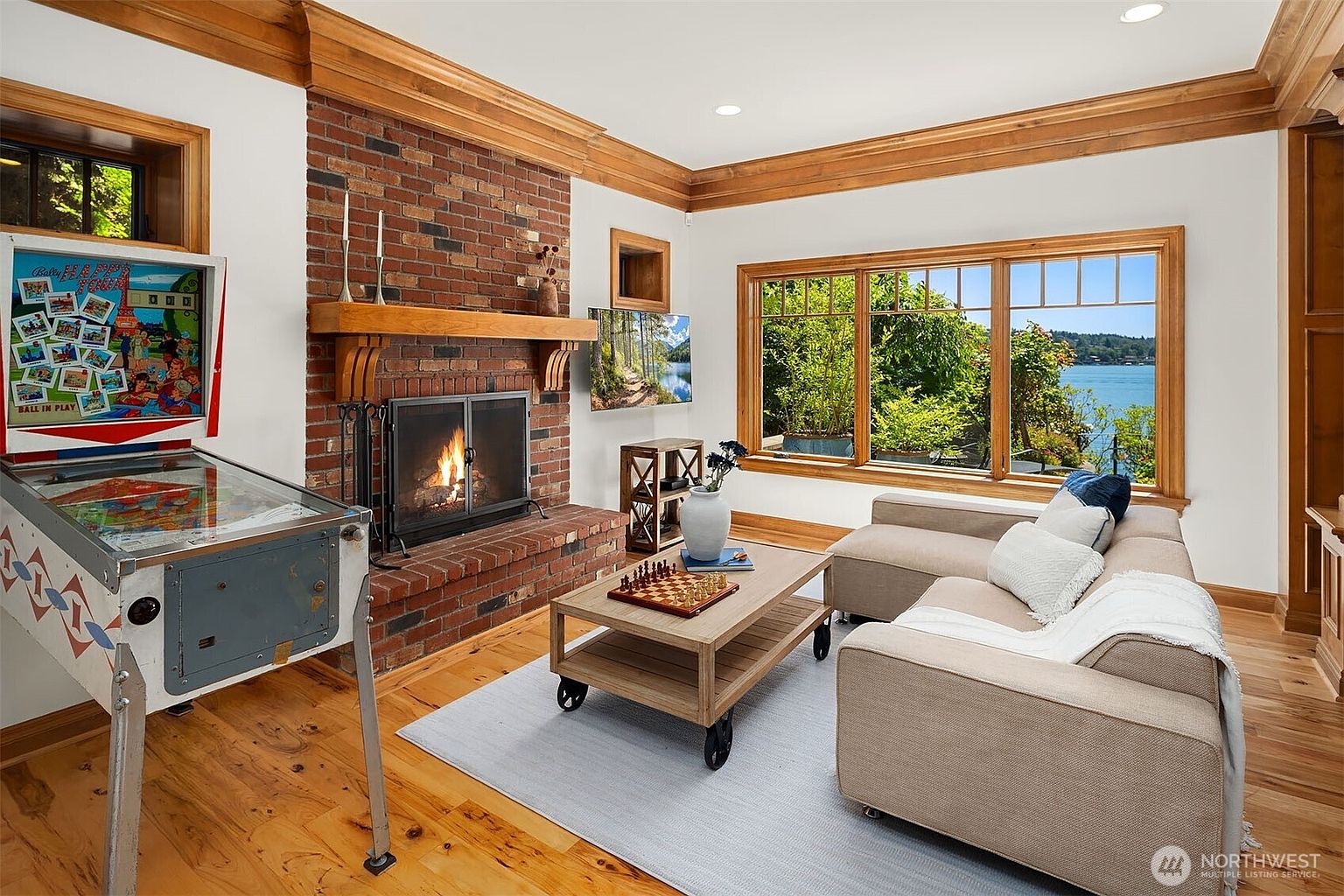 This cozy living room features a classic brick fireplace as its focal point, complemented by warm wood trim and hardwood flooring. A comfortable sectional sofa faces the fireplace and a large window that provides a stunning, panoramic view of the lake. The space is uniquely accented with a vintage pinball machine, adding a playful touch to the traditional, inviting atmosphere.