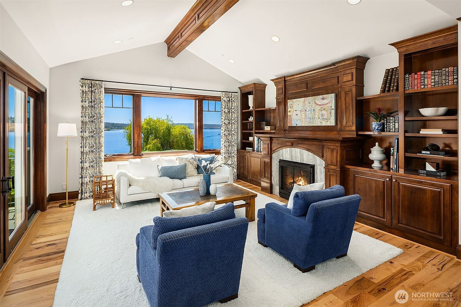 This elegant living room features a vaulted ceiling with exposed wooden beams and a large picture window offering a stunning view of the lake. The space is anchored by a cozy fireplace with a custom wood mantel and built-in bookshelves, complemented by a plush white sofa and two deep blue armchairs. The warm hardwood floors and soft area rug create a sophisticated yet inviting atmosphere perfect for relaxation.