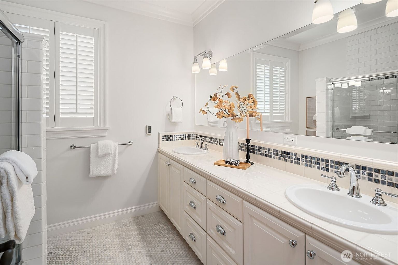 This bright and spacious primary bathroom features a long double vanity with white cabinetry, tiled countertops, and a decorative mosaic backsplash. A large wall-to-wall mirror reflects the room, enhancing the sense of space, while a glass-enclosed shower is visible on the left. The room is finished with classic white shutters and a light-colored hexagonal tile floor, creating a clean and traditional aesthetic.
