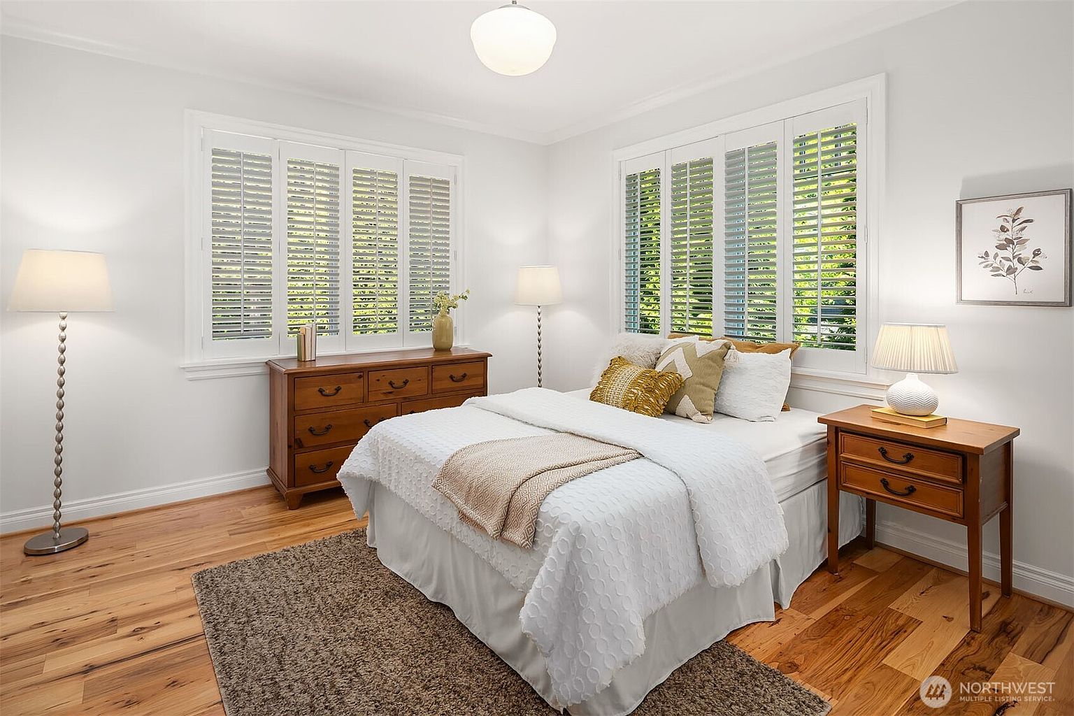 This bright and inviting bedroom features a queen-sized bed dressed in white linens with textured accent pillows, centered between two wooden nightstands. The room is accented by a warm-toned wooden dresser, plantation shutters on the windows, and a plush area rug over hardwood flooring. The overall aesthetic is clean, cozy, and traditional, creating a peaceful retreat.