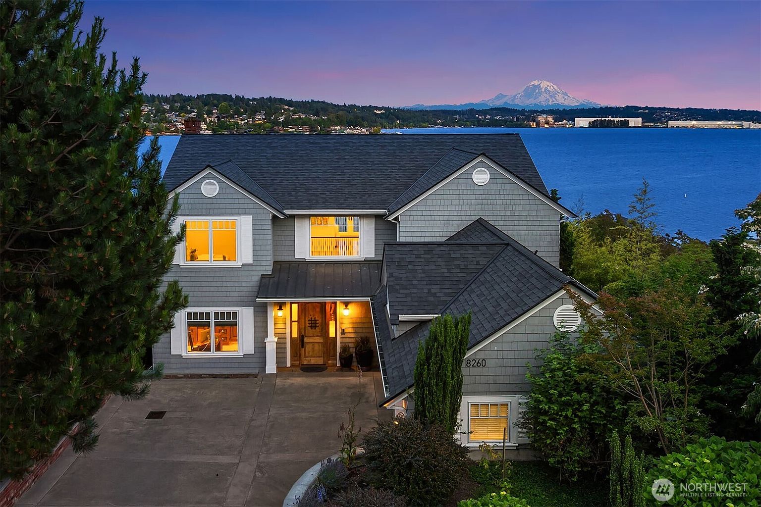 This stunning two-story waterfront home features classic shingle-style siding and a multi-gabled roofline, set against a breathtaking backdrop of a lake and a snow-capped mountain at dusk. The front entrance is warmly lit, welcoming visitors to a well-maintained driveway and landscaped yard. The composition captures the serene, high-end atmosphere of a luxury property with a prime scenic location.