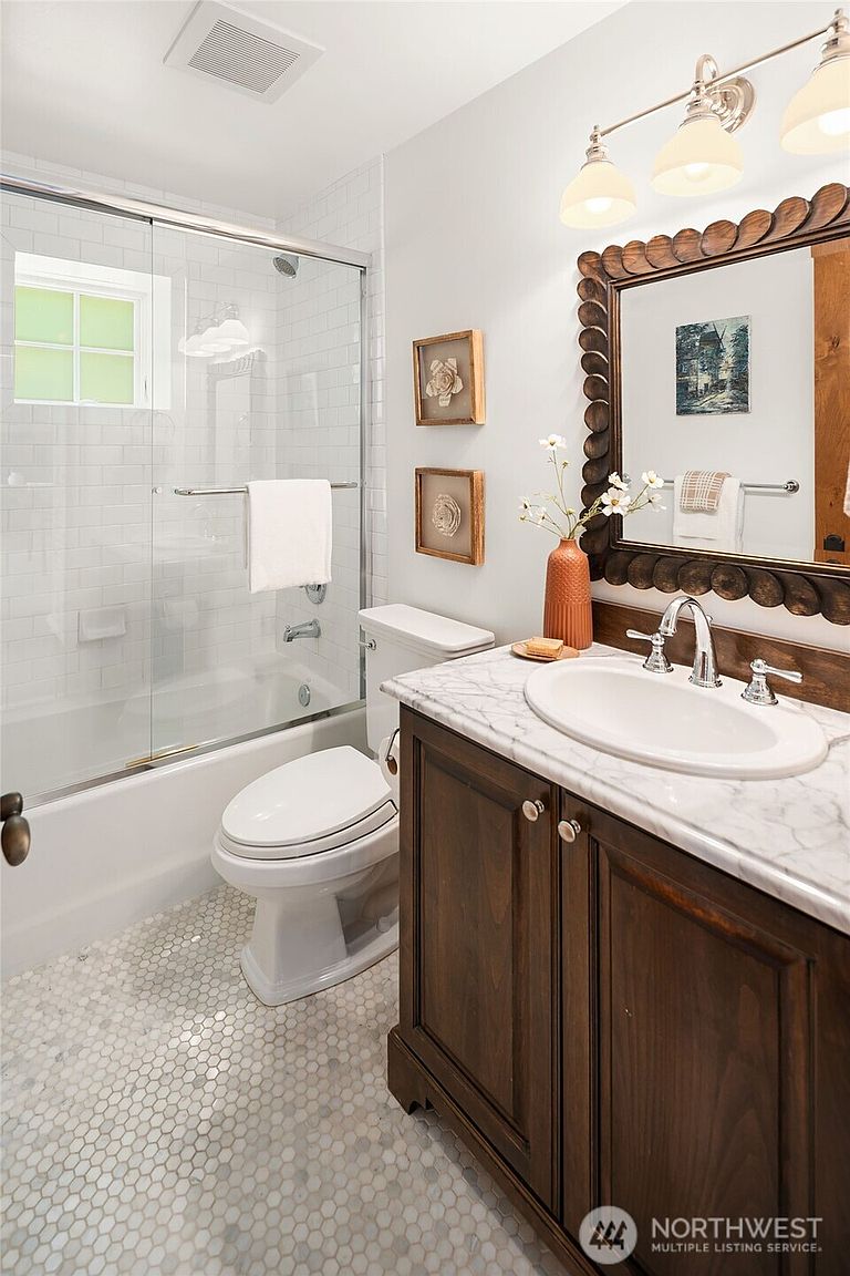 This well-appointed bathroom features a dark wood vanity topped with a white marble countertop and an oval undermount sink. A decorative, scalloped-edge mirror hangs above the vanity, complemented by a three-light fixture, while the shower area includes a glass-enclosed tub with white subway tile surround. The space is finished with classic hexagonal floor tiles and neutral wall decor, creating a clean and inviting aesthetic.