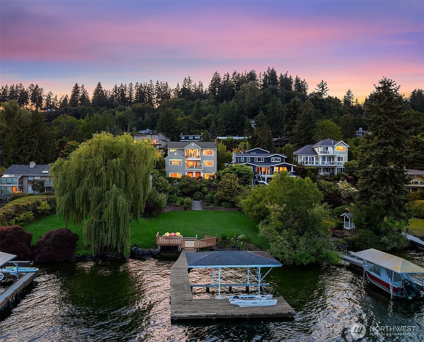 This stunning aerial view captures a multi-story waterfront residence at sunset, showcasing its expansive green lawn leading down to a private dock and boat lift. The home features multiple balconies and large windows that reflect the warm, vibrant colors of the twilight sky. Surrounded by lush trees and neighboring properties, the setting offers a serene and picturesque lakeside lifestyle.
