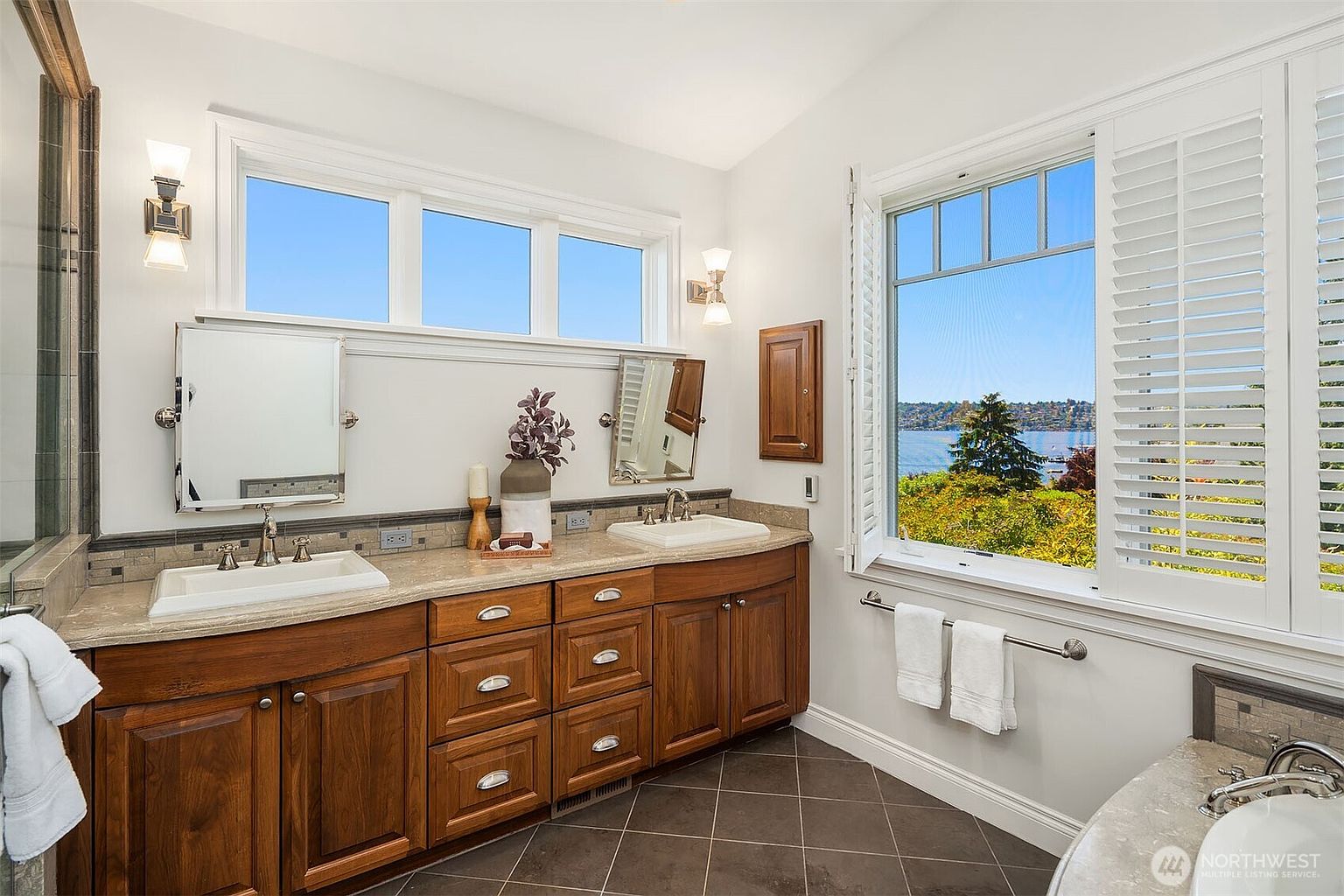 This elegant primary bathroom features a double vanity with rich wood cabinetry and a neutral stone countertop, complemented by dark tile flooring. The space is brightened by natural light from a large window offering a scenic water view and a row of transom windows above the mirrors. The overall aesthetic is sophisticated and serene, highlighting a high-end, well-maintained design.