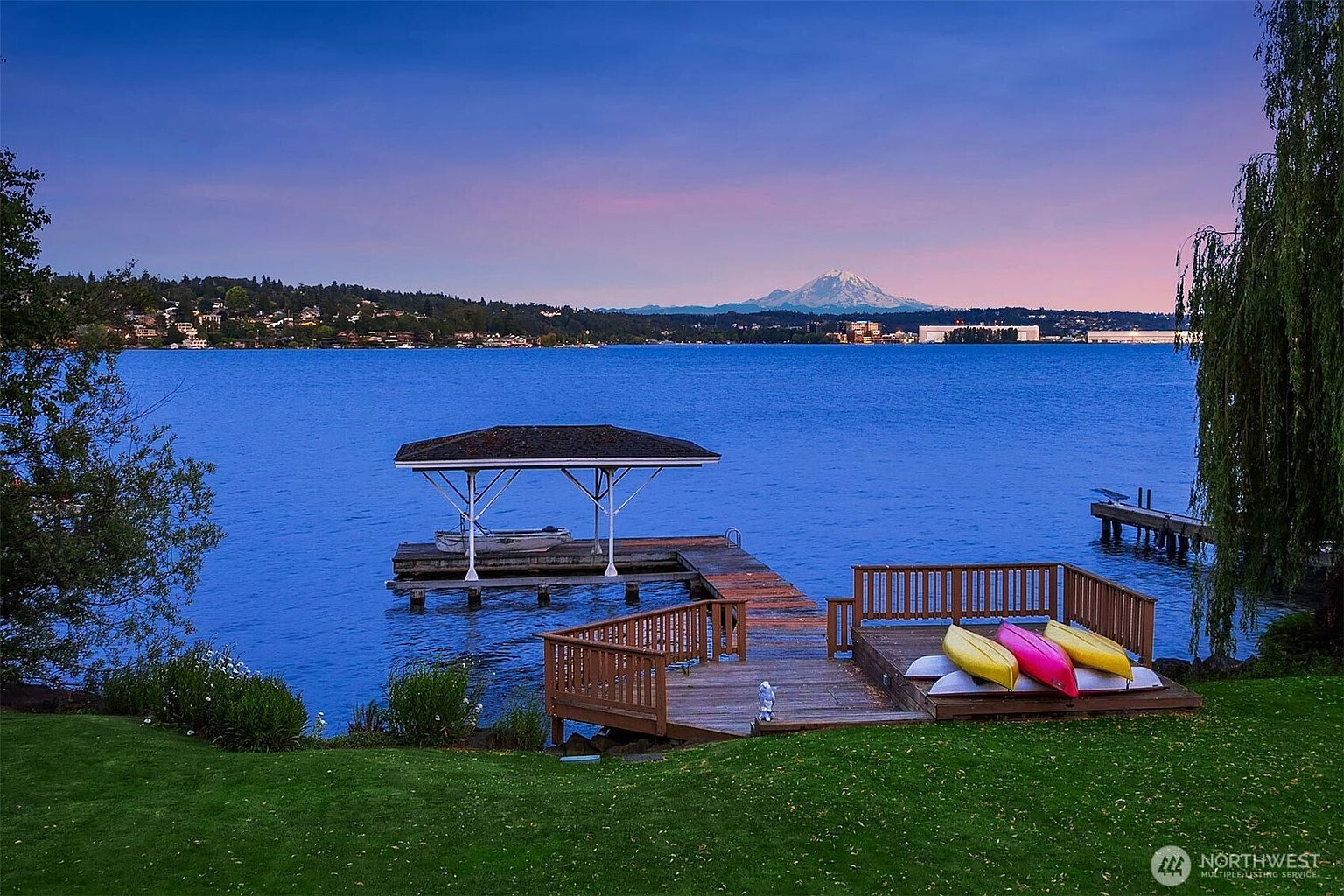 This scenic waterfront view features a wooden deck extending over a calm lake, complete with a covered boat slip and a trio of colorful kayaks resting on the dock. In the background, the majestic, snow-capped Mount Rainier rises above the horizon under a soft, twilight-hued sky. The perspective is captured from the lush green lawn, emphasizing the serene and picturesque lifestyle offered by this lakeside property.