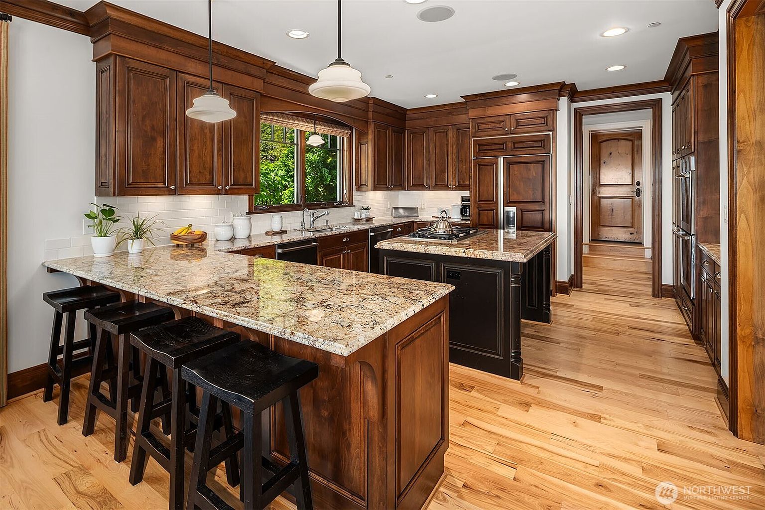 This spacious, traditional-style kitchen features rich, dark-stained wood cabinetry paired with elegant granite countertops and a large central island. The room is well-lit by pendant lights and recessed lighting, highlighting the warm hardwood floors and the seamless transition to an adjacent hallway. The overall impression is one of sophisticated warmth and functional luxury, perfect for both cooking and casual dining.