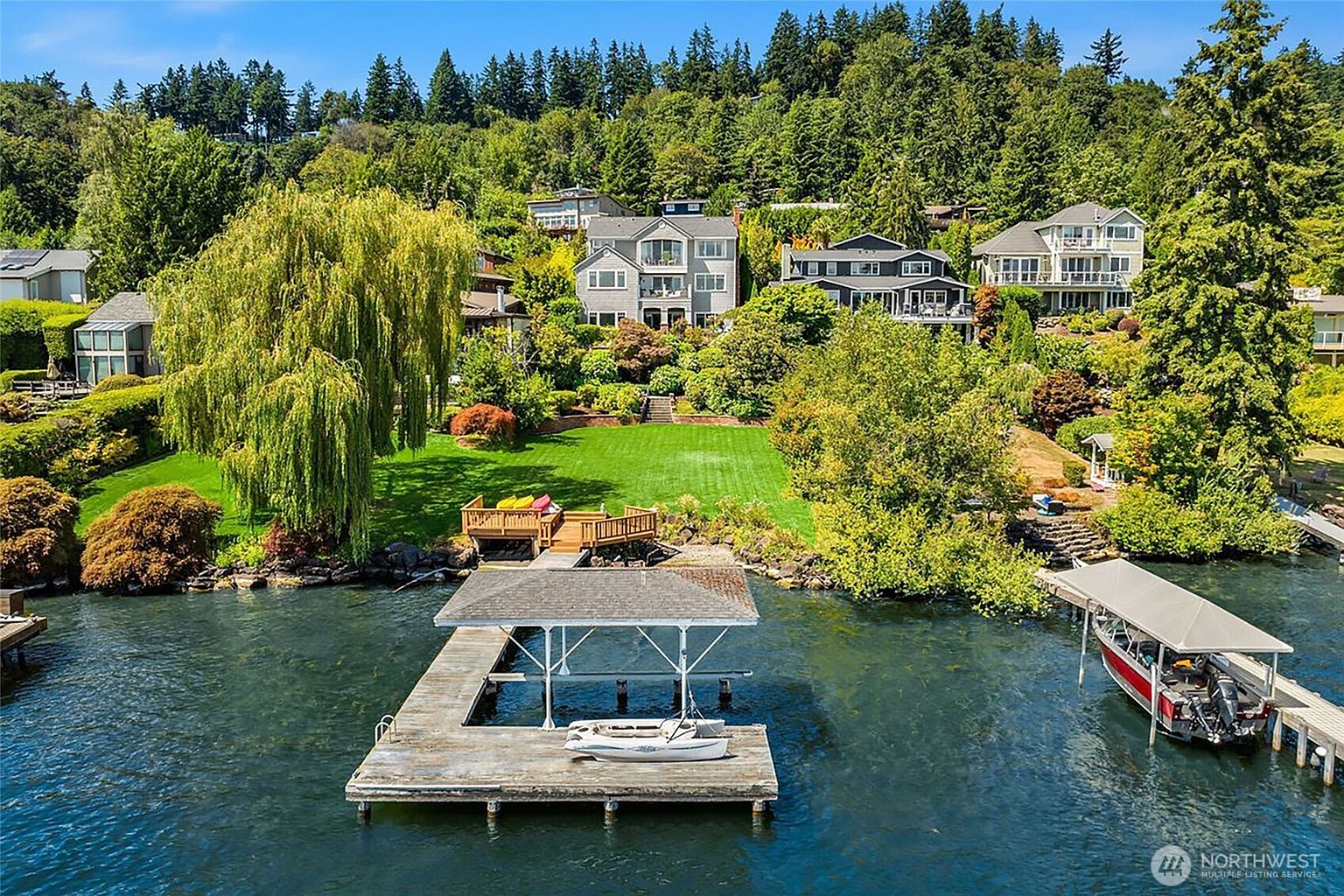 This aerial view captures a stunning waterfront property featuring a lush, expansive green lawn that slopes gently down to a private wooden dock and boat lift on the lake. The scene is framed by mature trees and tiered landscaping, with several multi-story residential homes nestled into the hillside in the background. The perspective highlights the serene, private nature of the property and its direct access to the water, creating an inviting and luxurious atmosphere.