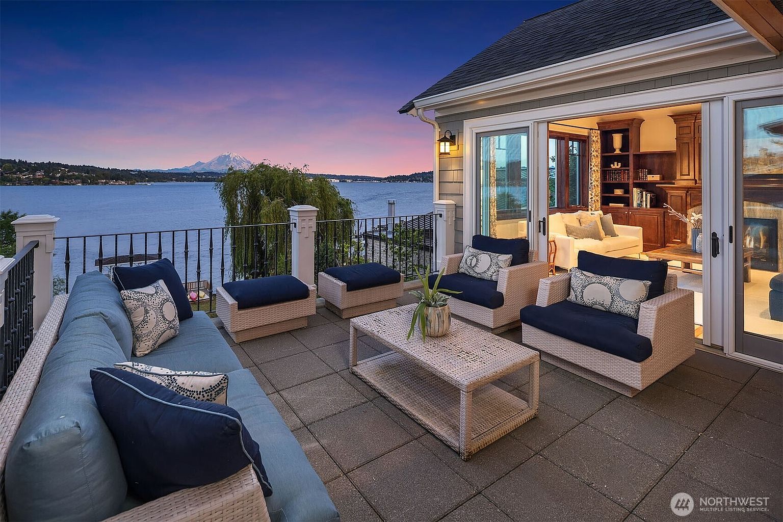 This stunning outdoor deck offers a luxurious lounge area with comfortable wicker furniture, plush blue cushions, and a central coffee table, all set against a breathtaking panoramic view of a lake and a snow-capped mountain at sunset. The space seamlessly connects to an interior living room through large glass sliding doors, creating an ideal indoor-outdoor flow for entertaining. The elegant iron railing and sophisticated lighting fixtures enhance the upscale, serene atmosphere of this waterfront property.