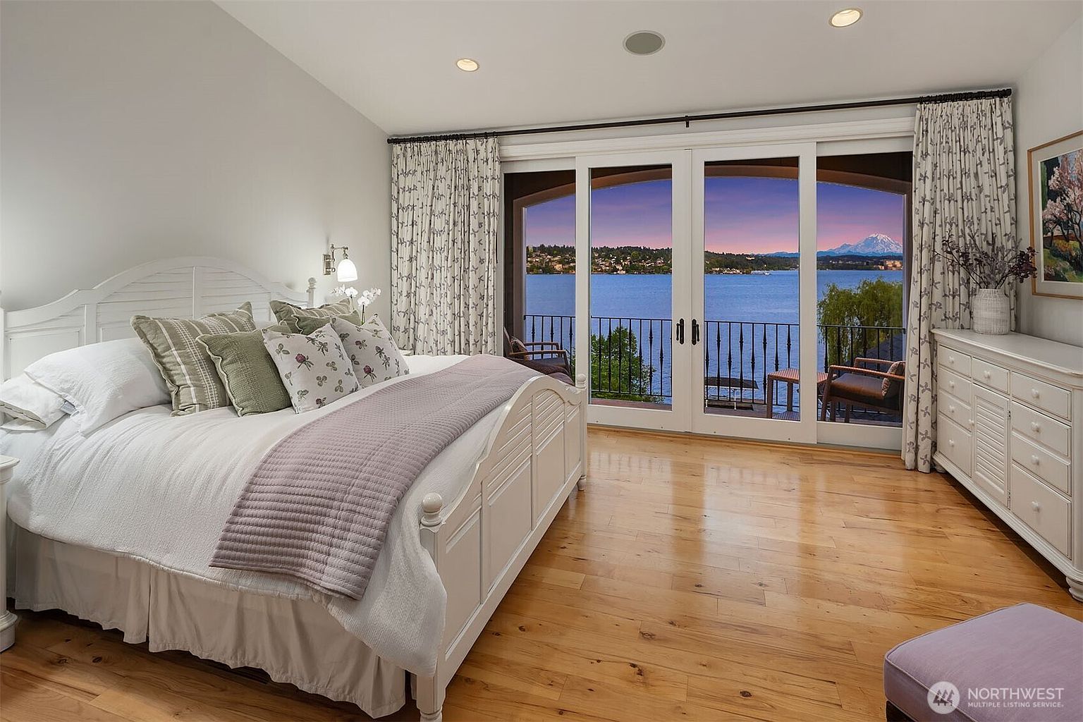 This elegant primary bedroom features a white wooden bed frame, a plush white duvet, and a soft lavender throw blanket, creating a serene and inviting atmosphere. The room is anchored by light hardwood floors and opens up to a private balcony through large glass sliding doors that frame a stunning, panoramic view of a lake and a distant snow-capped mountain. The space is complemented by a white dresser and patterned curtains, emphasizing a bright, airy, and luxurious aesthetic.