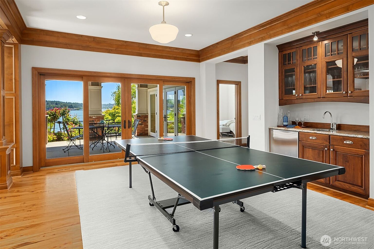 This spacious game room features a central ping pong table set atop a large area rug, creating a dedicated recreational space. To the right, a built-in wet bar with wooden cabinetry and a stainless steel under-counter refrigerator adds convenience, while large sliding glass doors on the left provide a seamless transition to an outdoor patio with scenic water views. The room is finished with warm wood trim and crown molding, offering a sophisticated yet relaxed atmosphere.