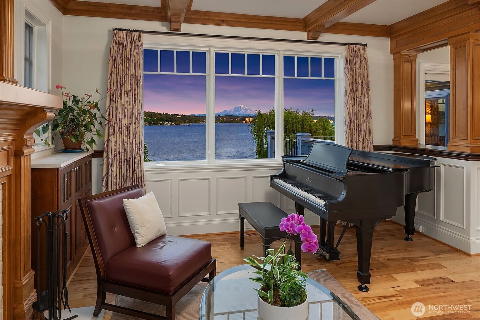 This elegant living space features a grand piano positioned in front of a large window that frames a breathtaking view of a lake and a snow-capped mountain at sunset. The room is appointed with rich wood trim, a leather armchair with a white accent pillow, and a glass-topped coffee table adorned with a vibrant orchid. The combination of classic musical elements and stunning natural scenery creates a sophisticated and serene atmosphere.