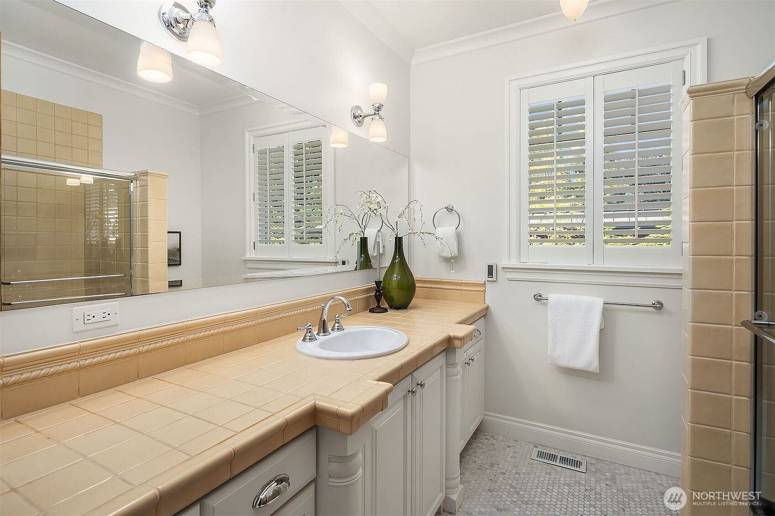 This bright and spacious bathroom features a large vanity with tan tile countertops and a white undermount sink, complemented by white cabinetry. A expansive wall-to-wall mirror reflects the room, while plantation shutters on the window allow for natural light. The space is finished with a classic mosaic tile floor and elegant light fixtures, creating a clean and functional aesthetic.