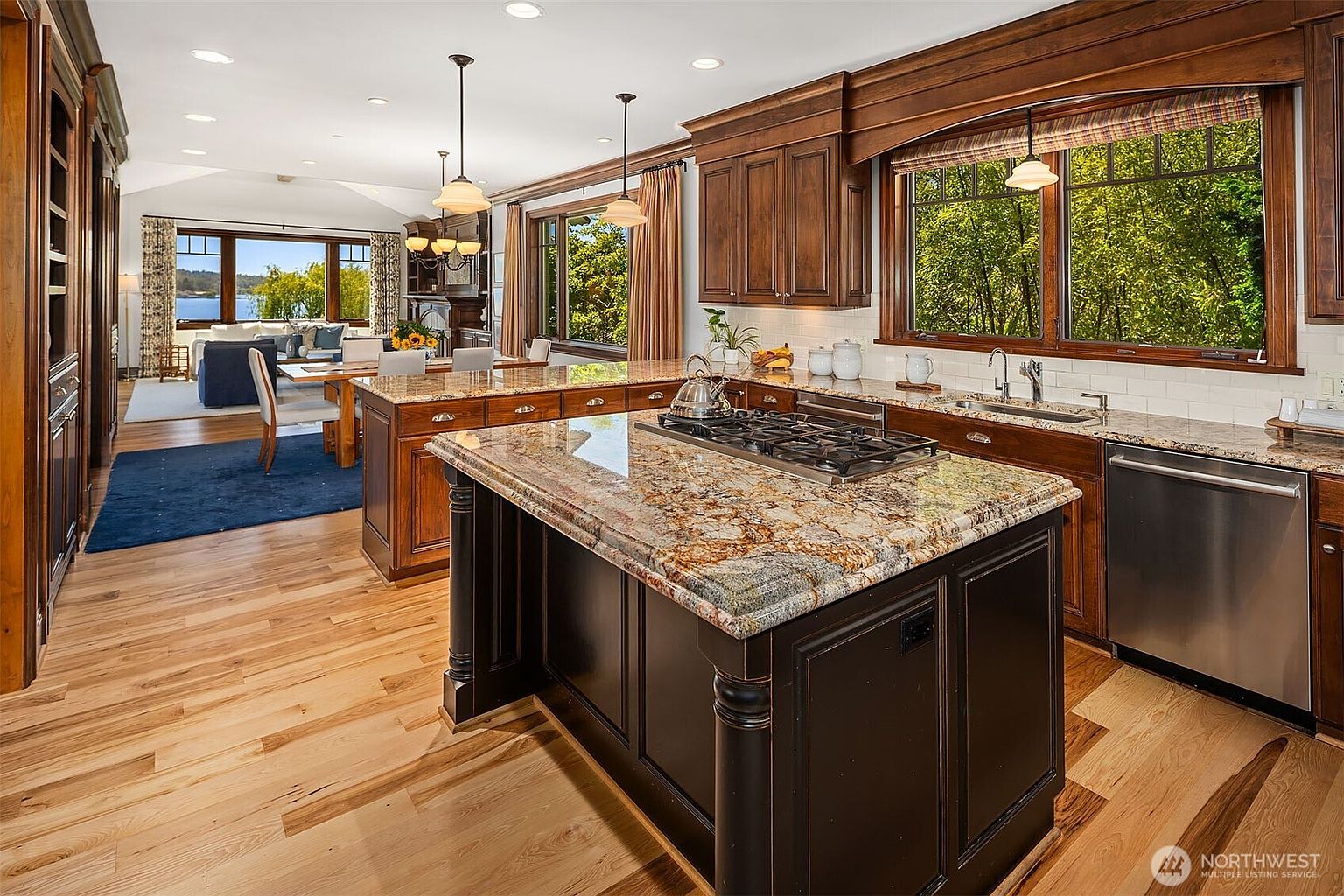 This elegant, traditional-style kitchen features rich, dark-stained wood cabinetry and a large central island topped with intricate, multi-toned granite countertops. The space is well-lit by recessed lighting and pendant fixtures, with a seamless transition into an open-concept dining and living area that overlooks a scenic waterfront view. The warm hardwood flooring and high-end stainless steel appliances create a sophisticated and inviting atmosphere perfect for both cooking and entertaining.