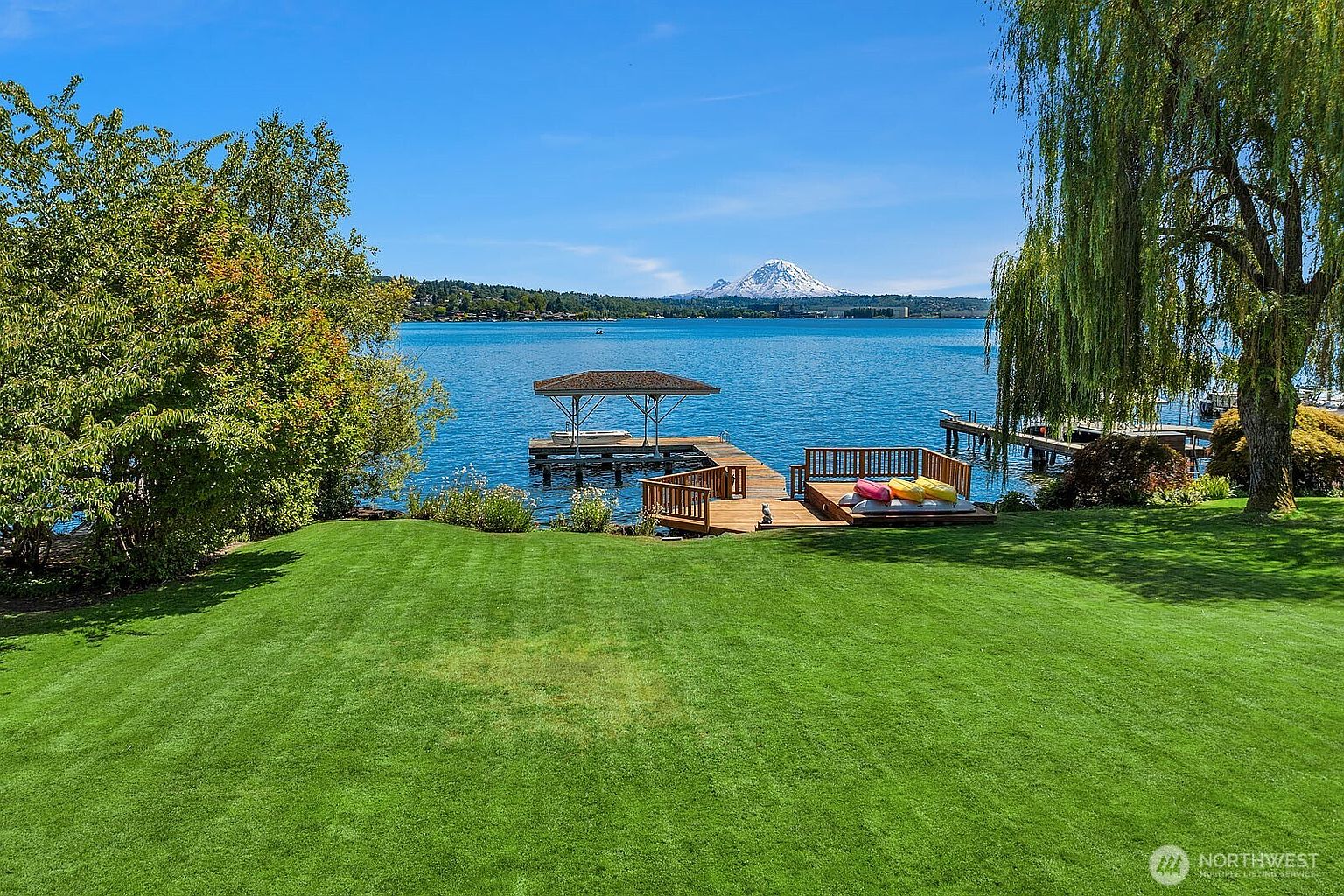 This stunning waterfront property features a meticulously manicured green lawn that leads directly to a private wooden dock and deck area on the lake. The scene is framed by lush trees and offers a breathtaking, unobstructed view of a snow-capped mountain in the distance. The perspective is a wide, eye-level shot that captures the serene transition from the grassy backyard to the expansive blue water.