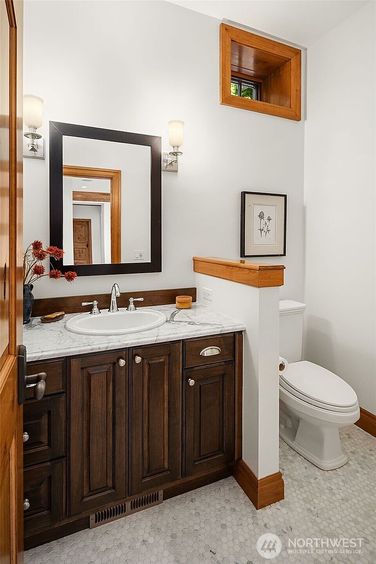 This elegant powder room features a rich, dark wood vanity topped with a white marble countertop and an undermount sink. The space is accented by a framed mirror, a small wall-mounted picture, and a unique high-set wooden window, all set against a crisp white wall and classic hexagonal tile flooring. A half-wall partition provides privacy for the toilet, creating a functional and sophisticated layout.
