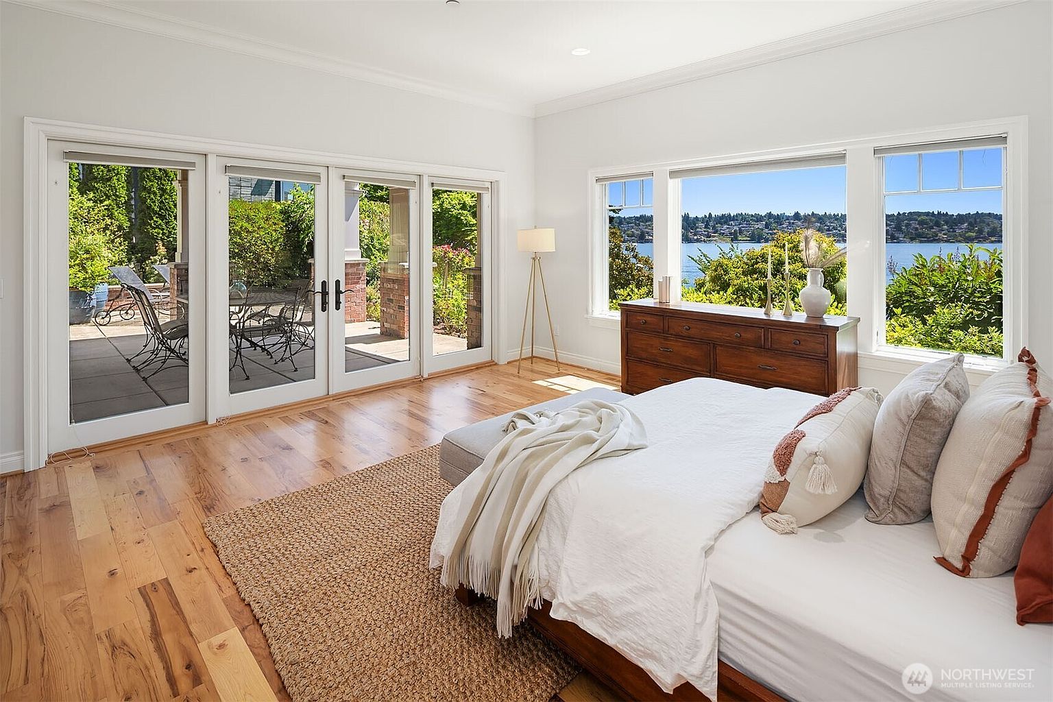 This bright and airy primary bedroom features light hardwood floors, a plush bed with neutral-toned bedding, and a large jute area rug. The room is flooded with natural light from expansive windows and glass doors that offer a stunning, unobstructed view of the lake and surrounding greenery. A wooden dresser sits beneath the windows, adding a touch of warmth to the clean, modern aesthetic.