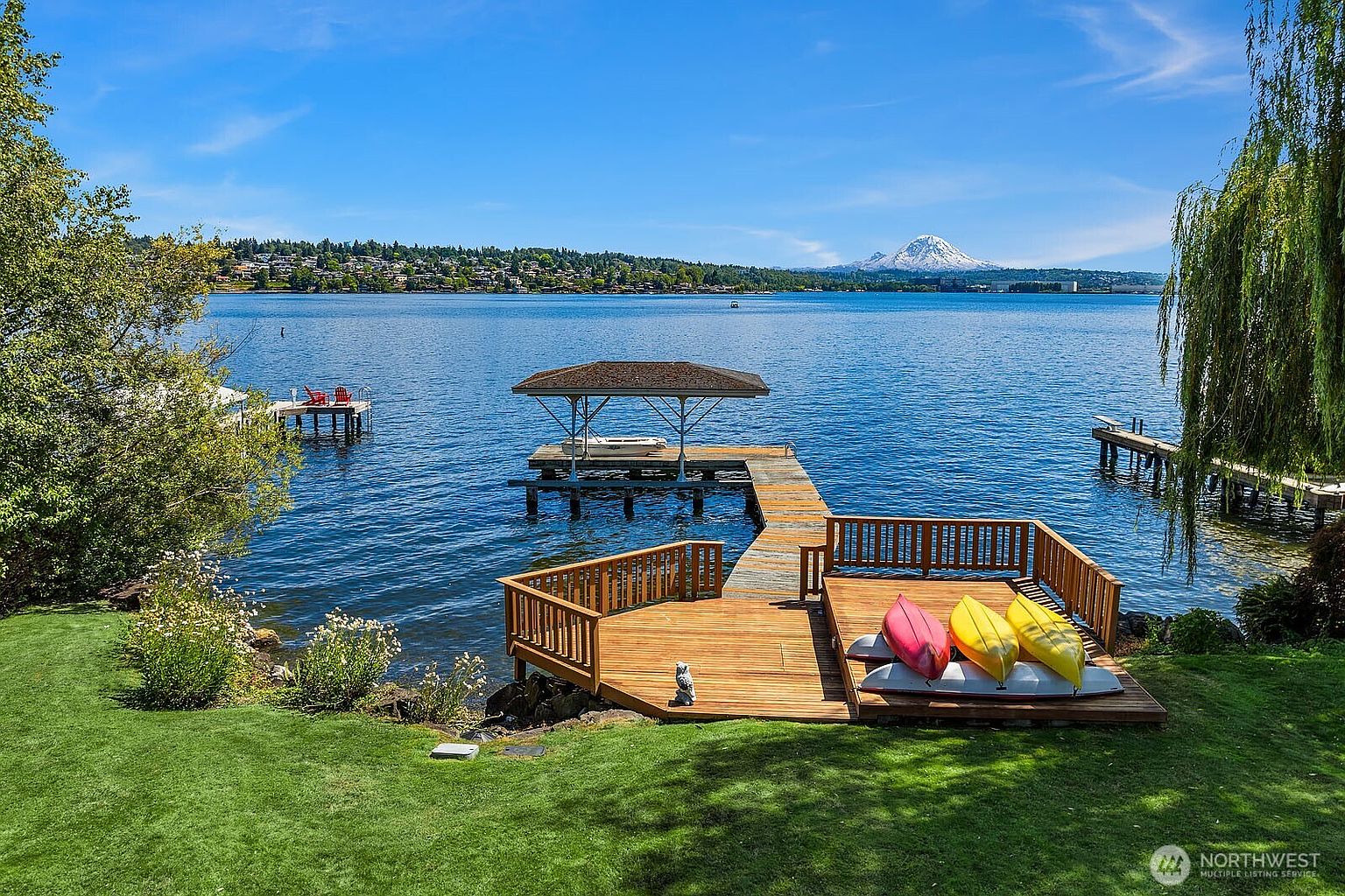 This scenic waterfront property features a beautifully maintained green lawn that transitions into a custom wooden dock extending over a calm lake. The dock includes a covered boat slip and a spacious deck area furnished with colorful kayaks, all set against a breathtaking backdrop of a snow-capped mountain. The perspective is a high-angle, wide shot that captures the serene atmosphere and the premium outdoor living space.