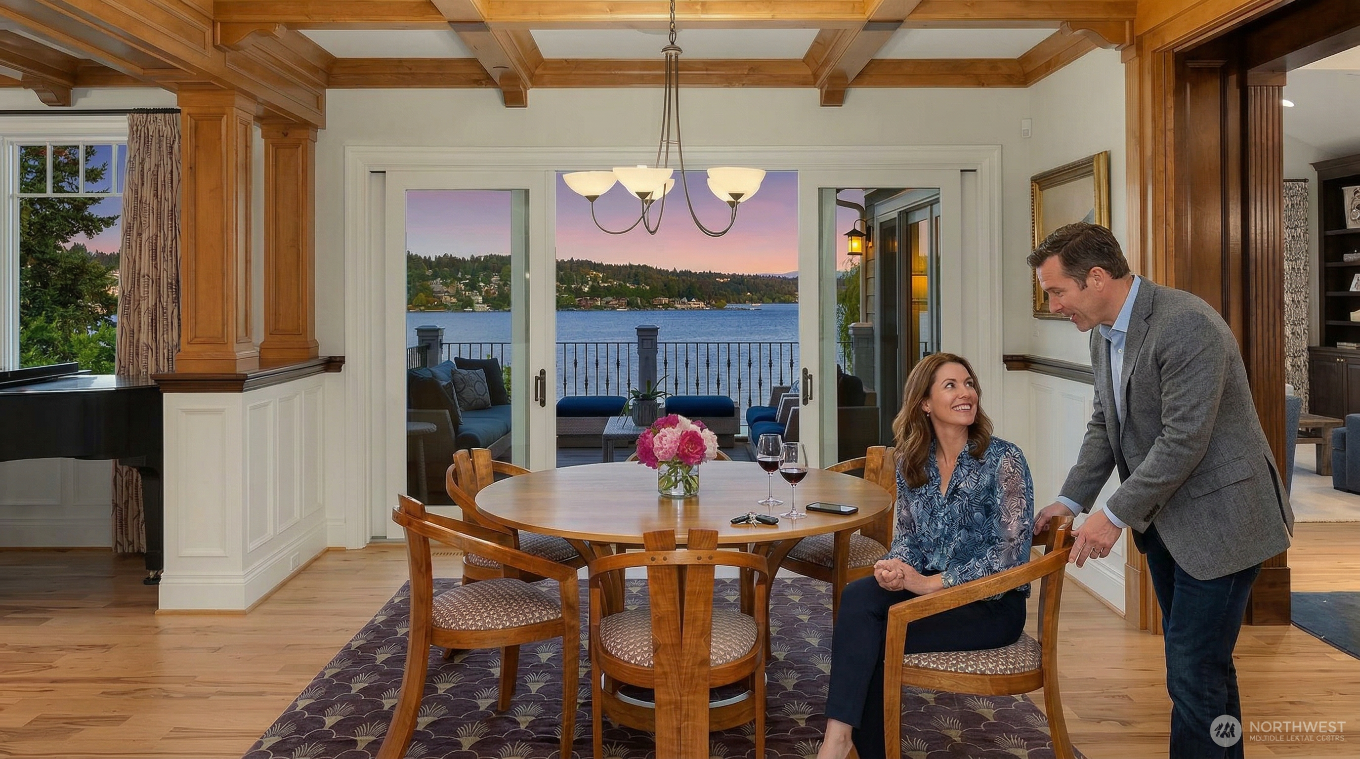 This elegant dining room features a warm, wood-toned aesthetic with coffered ceilings and rich hardwood flooring. A central round wooden table is surrounded by four upholstered chairs, all set atop a patterned area rug, while large sliding glass doors provide a stunning, unobstructed view of the lake and sunset. The space feels sophisticated and inviting, perfectly blending traditional architectural details with a seamless connection to the outdoor scenery.