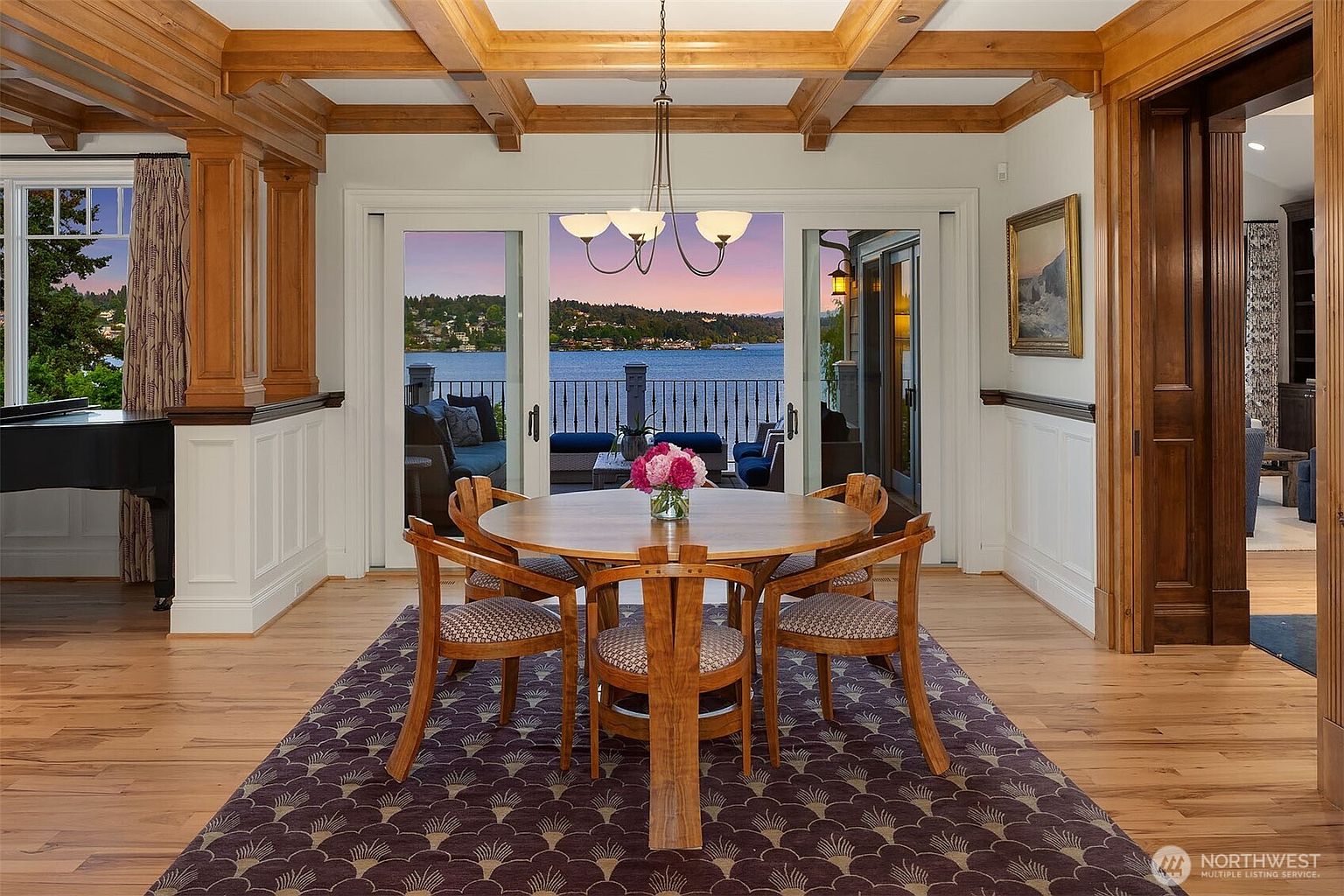 This elegant dining room features a warm, wood-toned aesthetic with coffered ceilings and rich hardwood flooring. A central round wooden table is surrounded by four upholstered chairs, all set atop a patterned area rug, while large sliding glass doors provide a stunning, unobstructed view of the lake and sunset. The space feels sophisticated and inviting, perfectly blending traditional architectural details with a seamless connection to the outdoor scenery.
