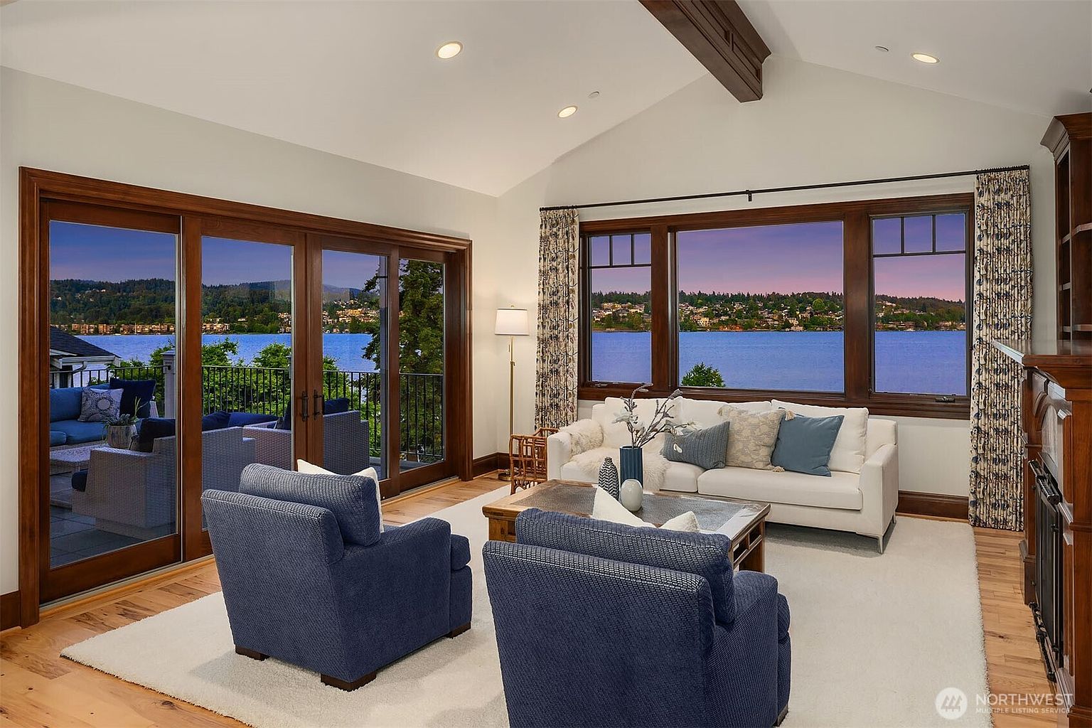 This elegant living room features a vaulted ceiling with exposed wooden beams and expansive windows that frame a stunning waterfront view. The space is furnished with a plush white sofa and two deep blue armchairs arranged around a rustic wooden coffee table on a soft area rug. The warm wood trim and natural light create a sophisticated, inviting atmosphere perfect for relaxing while overlooking the scenic landscape.