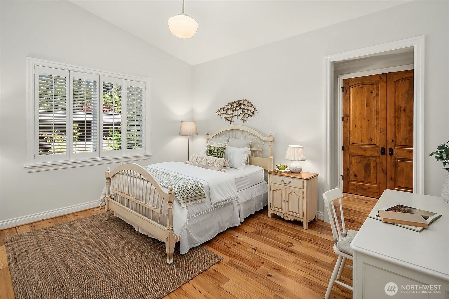 This bright and inviting guest bedroom features a classic white metal bed frame, light-colored hardwood flooring, and a neutral color palette. The room is accented by a decorative wall sculpture above the bed, plantation shutters on the window, and a rustic wooden door that adds a touch of warmth. A small desk and chair are positioned in the foreground, creating a functional space that feels both cozy and well-appointed.